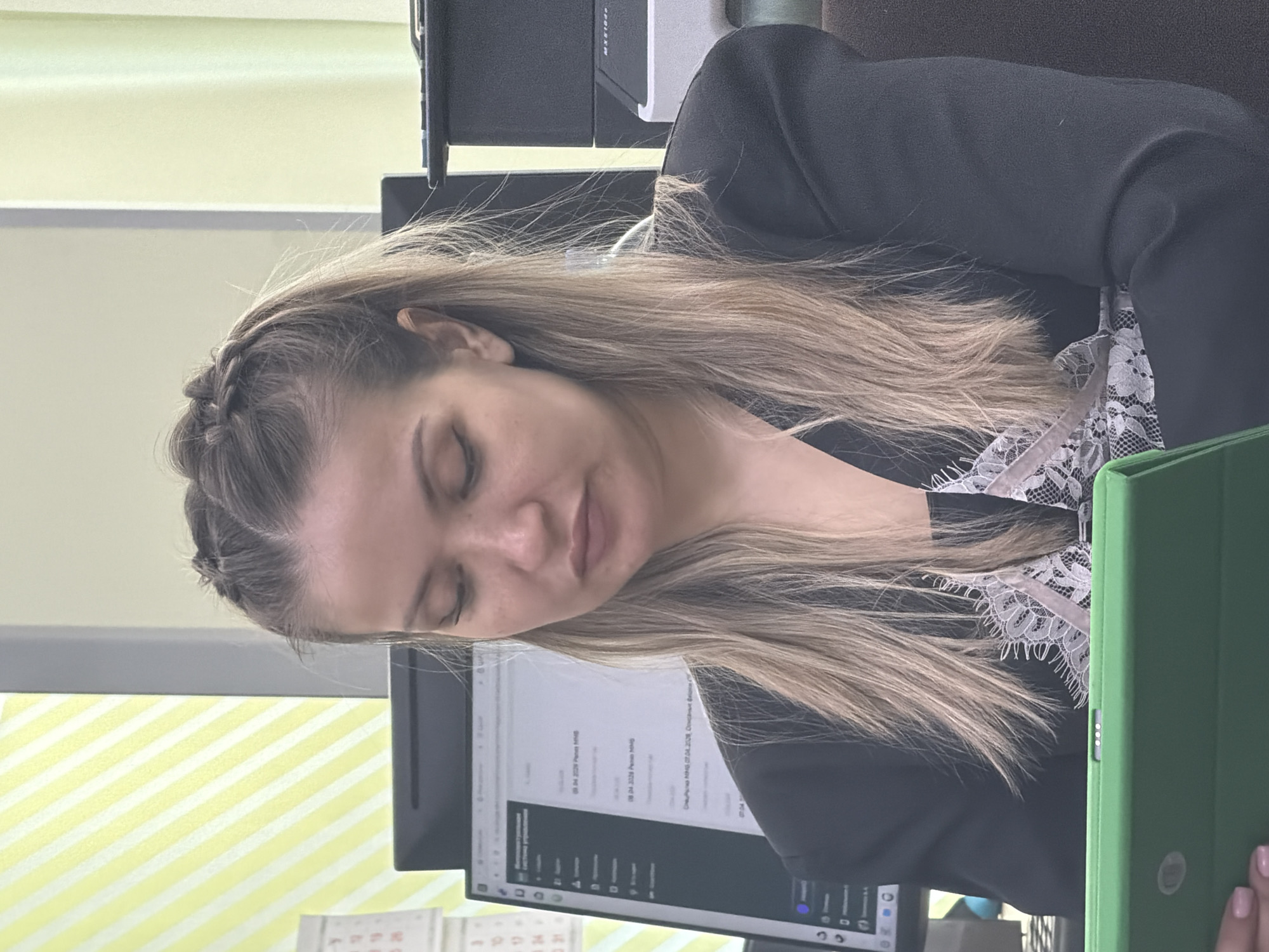 A woman with braided hair, wearing a black blazer over a white lace-trimmed top, is seated at a desk. A green tablet is held in her right hand. A computer monitor is visible in front of her, displaying what appears to be a work-related document. A light-colored striped wall is partially visible in the background. Based on the image details, the location could potentially be an office setting in Krasnoyarsk, Russia, considering the presence of a document with Russian language.