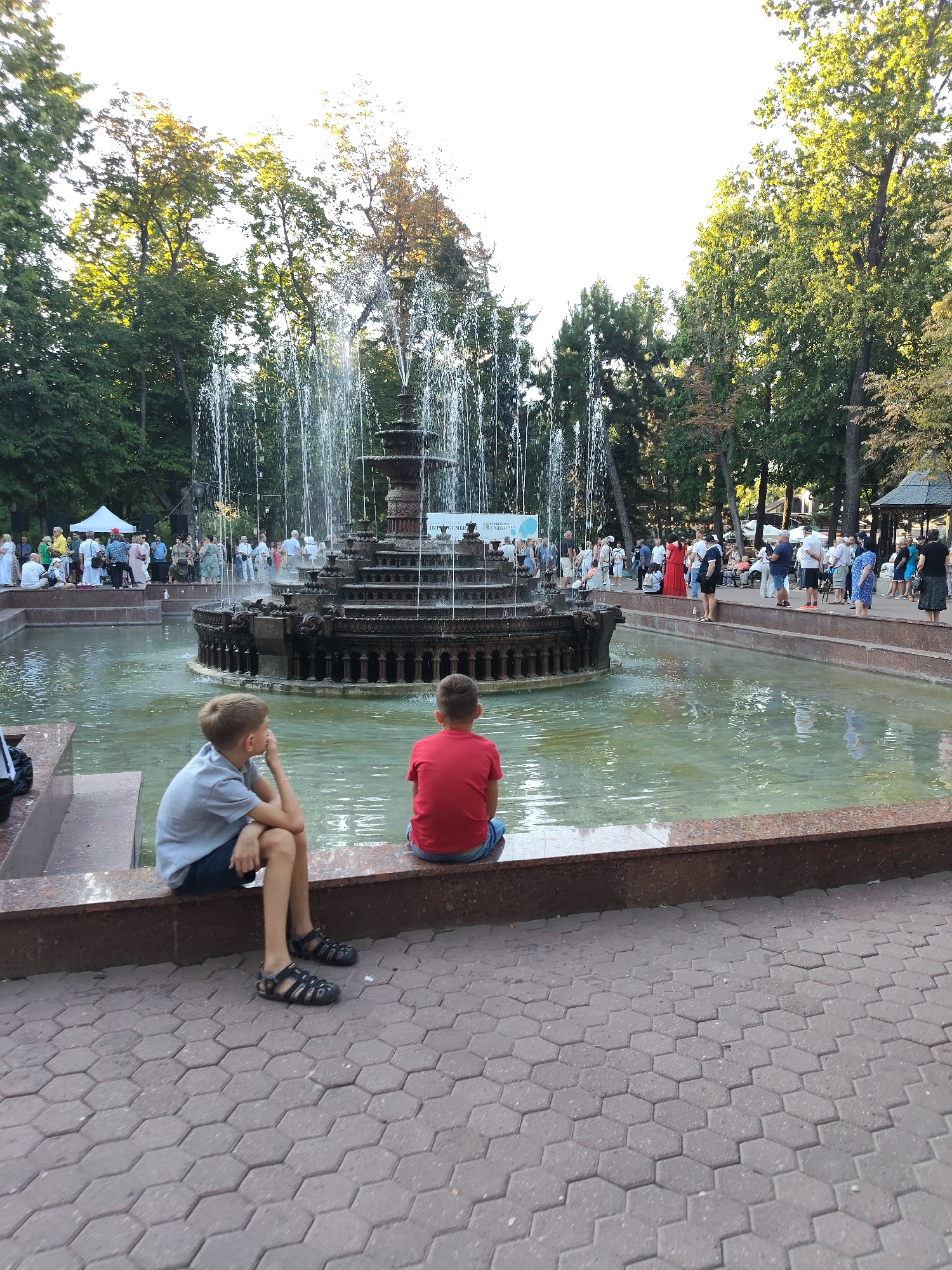 In Chișinău, Moldova, a public park scene unfolds around a large, multi-tiered fountain. The fountain features a central structure with cascading water jets, surrounded by a pool of greenish water. Two young boys are seated on the edge of the fountain's basin, facing away from the camera and towards the fountain. The boy on the left wears a light gray t-shirt and dark shorts, while the boy on the right is dressed in a red t-shirt and blue shorts.
Behind the boys and the fountain, a crowd of people are gathered, some standing and observing, others possibly engaged in conversation. The background is filled with lush green trees, suggesting a park setting. The ground in the foreground is paved with hexagonal stone tiles. The sky above is bright, indicating daytime. The overall atmosphere appears to be a relaxed gathering in an urban park.