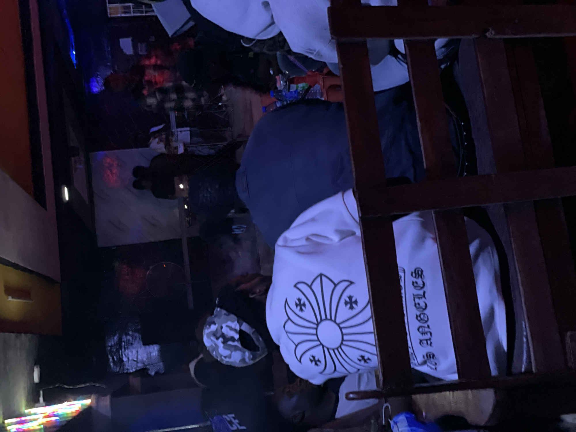In Durumi, Nigeria, a group of people are gathered indoors in what appears to be a dimly lit entertainment venue. The focus is on a person in the foreground wearing a white shirt with a large, intricate floral design featuring cross shapes on the back. Below this design, the words "Los Angeles" are visible in a gothic font. The person is seated at a wooden table, partially obscured by the wooden slatted backrest of a chair. To their left, another individual is visible from the shoulder up, looking to the side.
In the background, several other individuals are present, some of whom are obscured by darkness or other people. A group of people are standing near a stage or performance area, where musical equipment, including keyboards and a drum set, are set up. The environment is characterized by low lighting, with colorful neon lights illuminating the space. There are bottles of drinks and possibly a hookah pipe on a table in the middle ground, suggesting a social gathering or a place of leisure. The overall impression is of a lively evening event within a Nigerian city.
