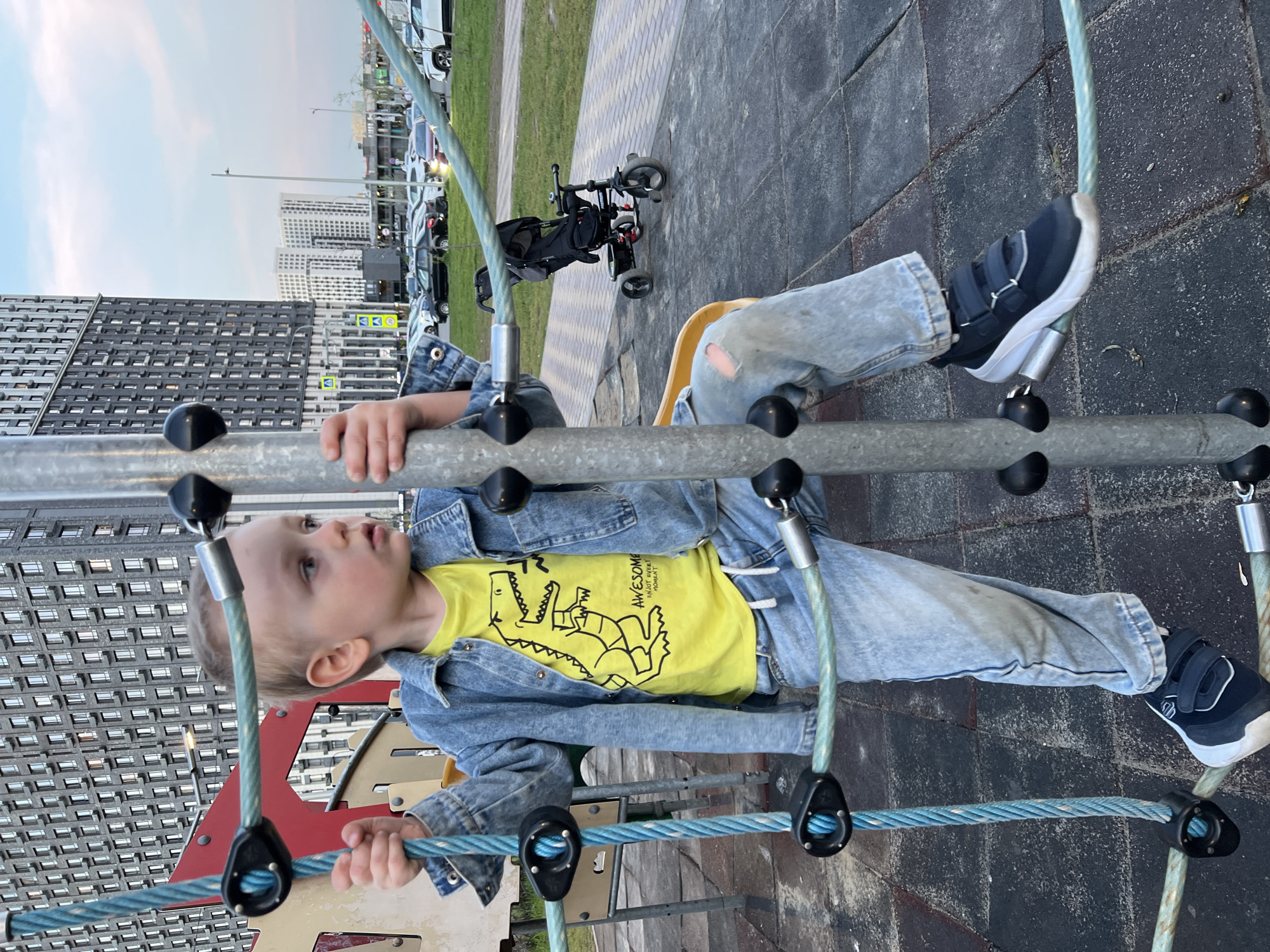 A young child, likely a boy, is on a playground structure composed of climbing ropes and metal bars with black spherical connectors. He's wearing a yellow t-shirt with a dinosaur graphic, a light blue denim jacket, and light blue jeans with a hole at the knee. He has dark blue and white sneakers on his feet. He is holding onto the metal bars.
In the background, tall apartment buildings typical of urban architecture are visible, indicating a city environment. A grassy area and a paved path are also present. In the background towards the right, there are cars parked, and an upright stroller can be identified.
The location is consistent with an urban residential area in the city of Mytishchi, Russia.