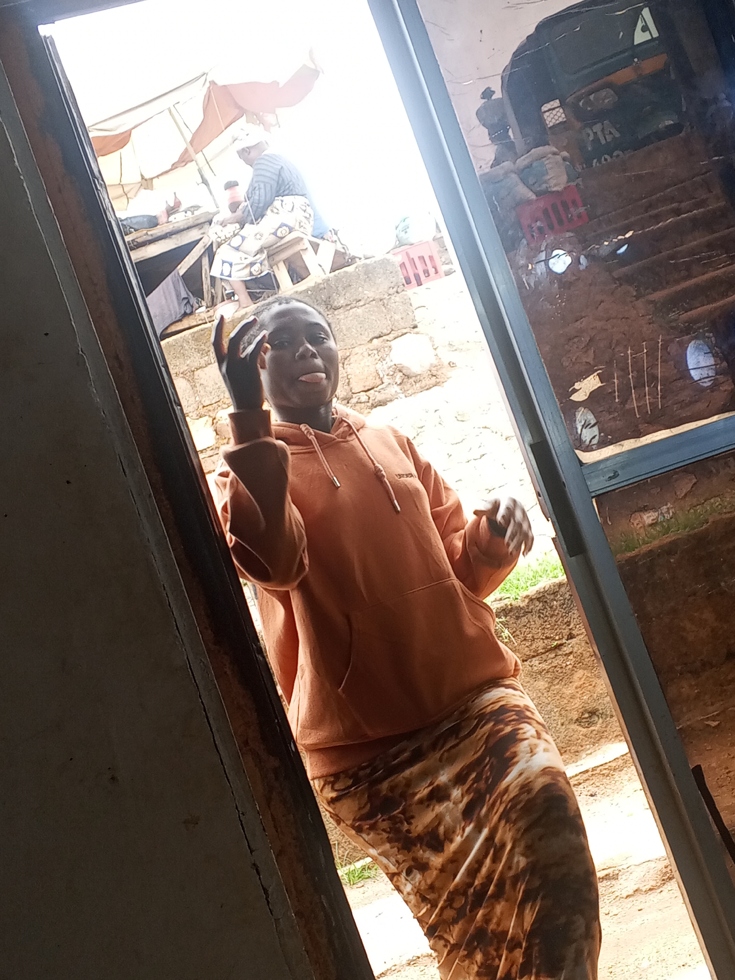 A young woman in Gwong, Nigeria, stands in a doorway. She wears an orange hoodie and a long, patterned skirt. She is sticking out her tongue and making a playful gesture with her hand. Through the doorway, there is a view of the street, including a table with people sitting under an umbrella and a motorized tricycle parked on a set of steps. The surrounding environment appears to be urban, with buildings and evidence of human activity visible.