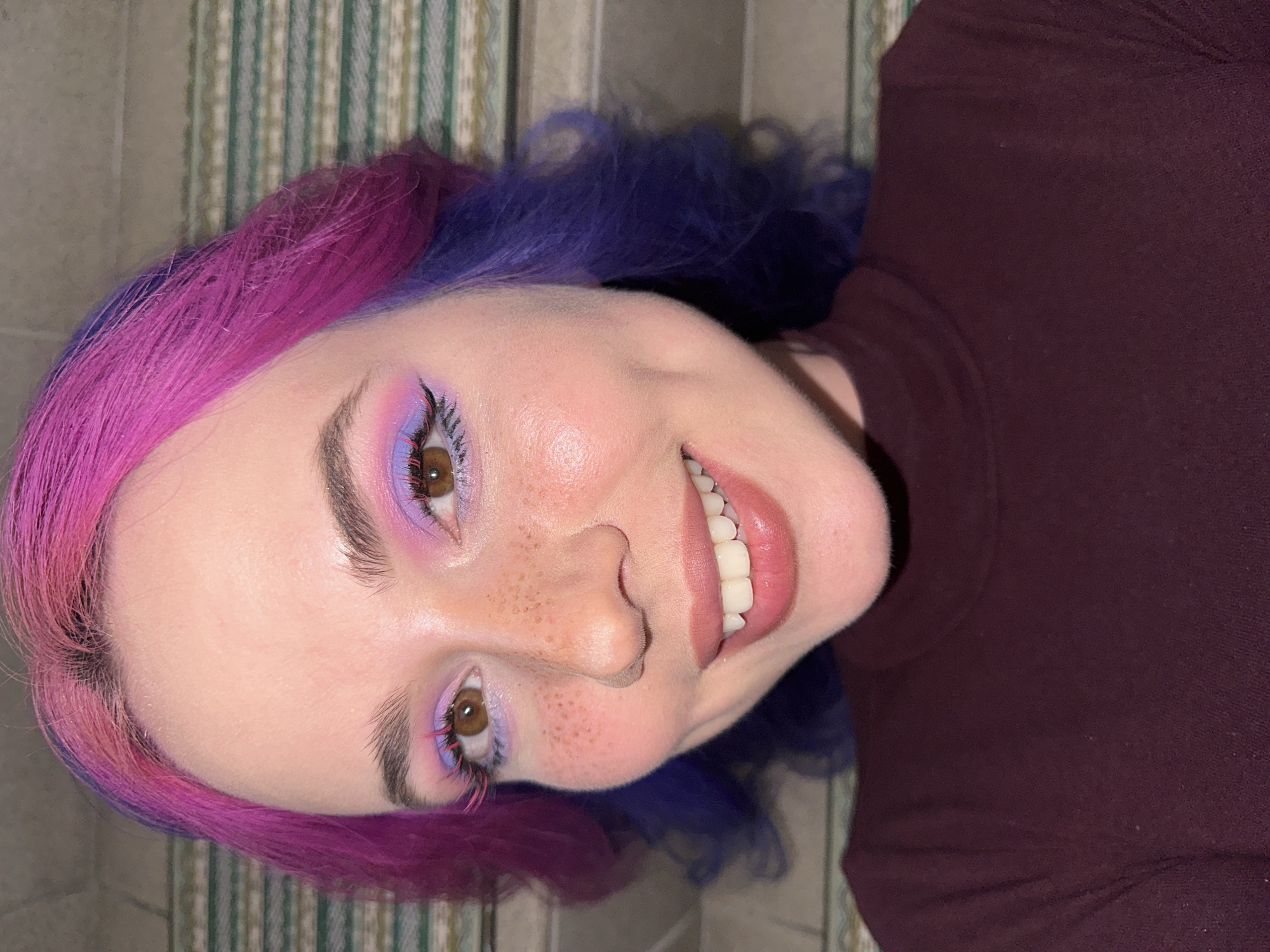 The image features a close-up portrait of a person, likely female, captured from the chest up with a joyful expression. They have distinctive split-dyed hair, with vibrant magenta-pink on the left side and a deep blue-purple hue on the right, styled with soft waves.
The person's brown eyes are adorned with elaborate and colorful makeup, featuring blended pink and purple eyeshadow, black eyeliner, and prominent bright pink artificial lower eyelashes. Freckles are visible across their nose and upper cheeks. A wide, open-mouthed smile reveals healthy white teeth and a small silver piercing on the tongue.
They are wearing a close-fitting, high-neck top in a dark reddish-brown (burgundy or maroon) color. The image is angled, suggesting the person might be lying down or tilting their head while looking directly at the camera.
The background consists of a vertical striped pattern, likely a wall or wallpaper, featuring alternating light green and off-white/beige stripes. A plain, light gray surface is partially visible on the far left. The lighting suggests an indoor setting. No text is visible in the image.