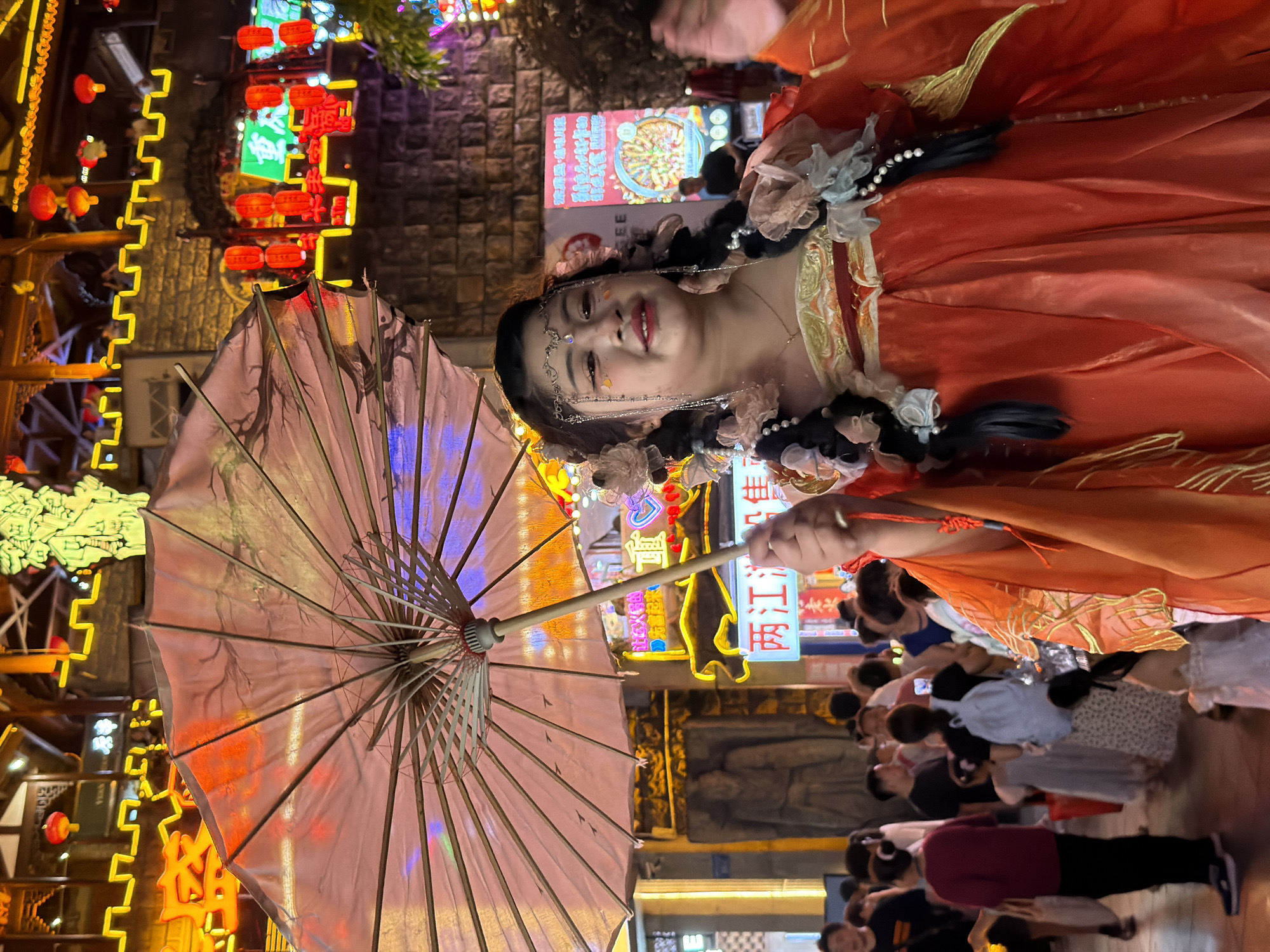In Yuzhong District, China, a young woman in a traditional orange dress adorned with floral and pearl accents is the central focus of the image. She holds a translucent umbrella with a pinkish hue, its spokes clearly visible against the vibrant, neon-lit background. Her hair is styled in dark braids decorated with ribbons and pearls, and she wears delicate jewelry on her forehead and ears, along with glittery eye makeup. She is smiling slightly, looking towards the right.
The background depicts a bustling street scene at night, illuminated by numerous red lanterns and bright neon signs, some in Chinese characters. The architecture suggests a historic or culturally themed area, with ornate structures and stone facades. In the middle ground and background, many people are visible, some appearing to be tourists or shoppers, with various casual attire. The overall atmosphere is lively and festive, typical of a night market or a cultural celebration.