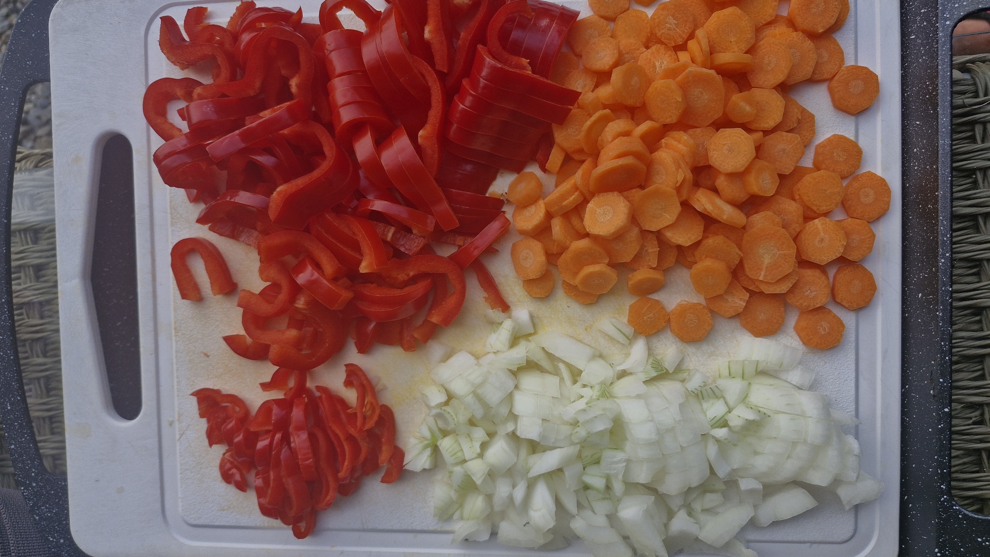 The image shows a close-up, top-down view of a white cutting board with chopped vegetables. On the left side, there are diced white onions. To the right of the onions, there are sliced rounds of bright orange carrots. The top and right portions of the cutting board are filled with sliced red bell peppers, some of which are cut into strips and others into crescent shapes. The cutting board is placed on a textured surface, possibly a placemat or table, with a dark rim along the top and bottom edges. The scene is well-lit, highlighting the vibrant colors of the vegetables. There is no indication of people, actions, or interactions. The location of Wattenwil, Switzerland, is not visually discernible from the image itself.