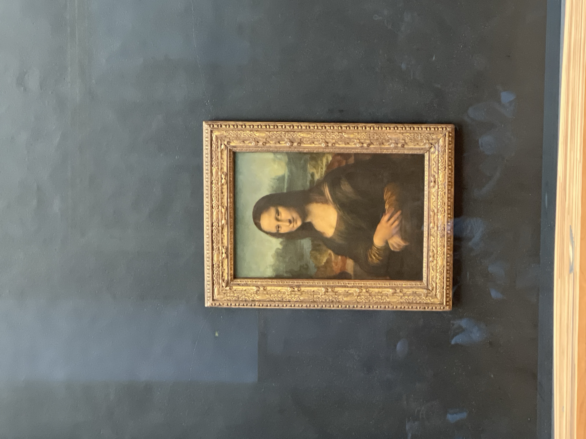 The original image is rotated 90 degrees clockwise, presenting a sideways view of the renowned Mona Lisa painting. The painting itself, a portrait of a woman with a gentle expression and folded hands set against a distant landscape, is housed in an ornate, gilded frame with intricate decorative patterns. It is displayed against a dark, possibly dark blue or charcoal grey, wall which shows signs of wear, including varying lighter smudges, dust, and faint marks, suggesting an aged surface. On the far right side of the image, a vertical section of a light-colored, possibly wooden, architectural feature, such as a door frame or paneling, is partially visible. The scene appears to be indoors under ample lighting. No specific activity or event is taking place other than the presentation of the artwork. There is no visible text in the image.