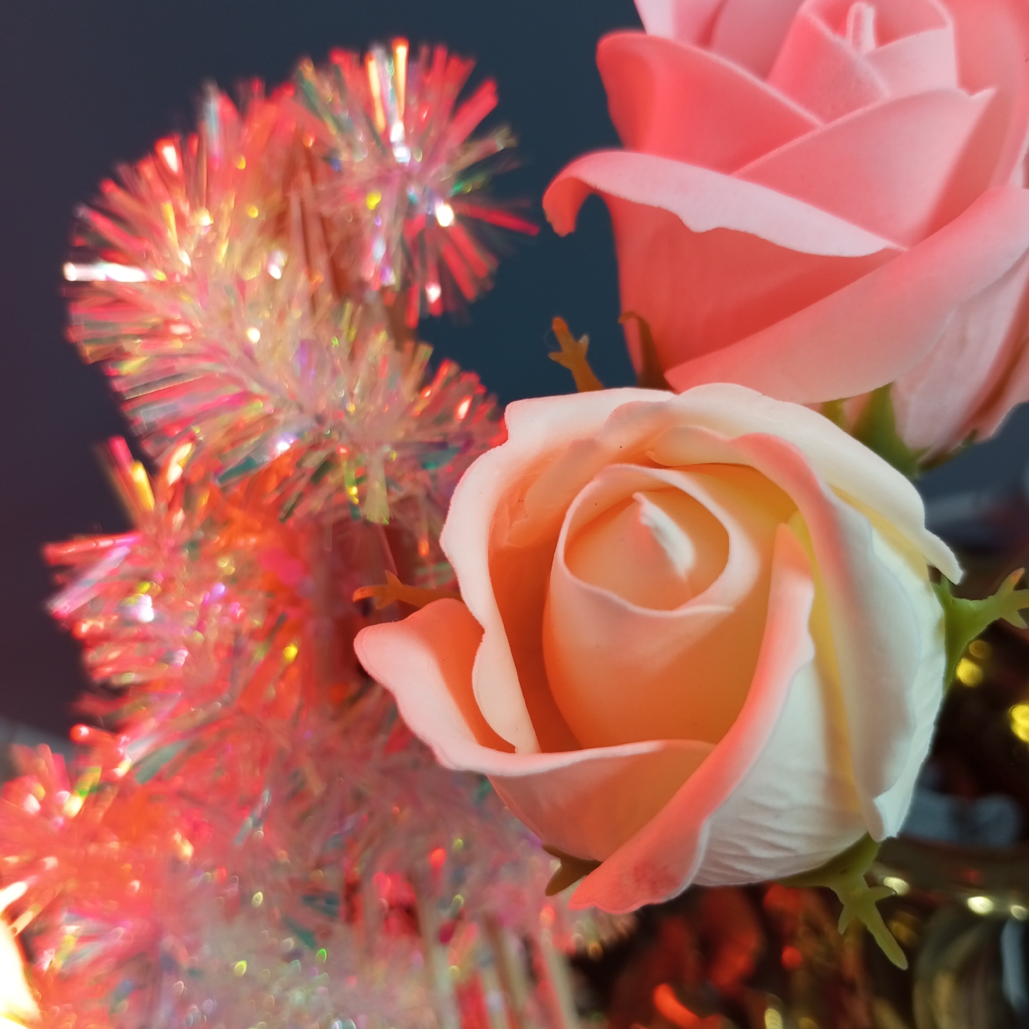The image shows two artificial roses, one peach and one pink, in the foreground. A shimmering, iridescent tinsel decoration is visible behind the roses, out of focus. The background appears to be a dark blue or gray color. The lighting is warm, casting a pinkish glow on the scene. The location in the city of Pervomaysky, Russia is not discernible from the image.