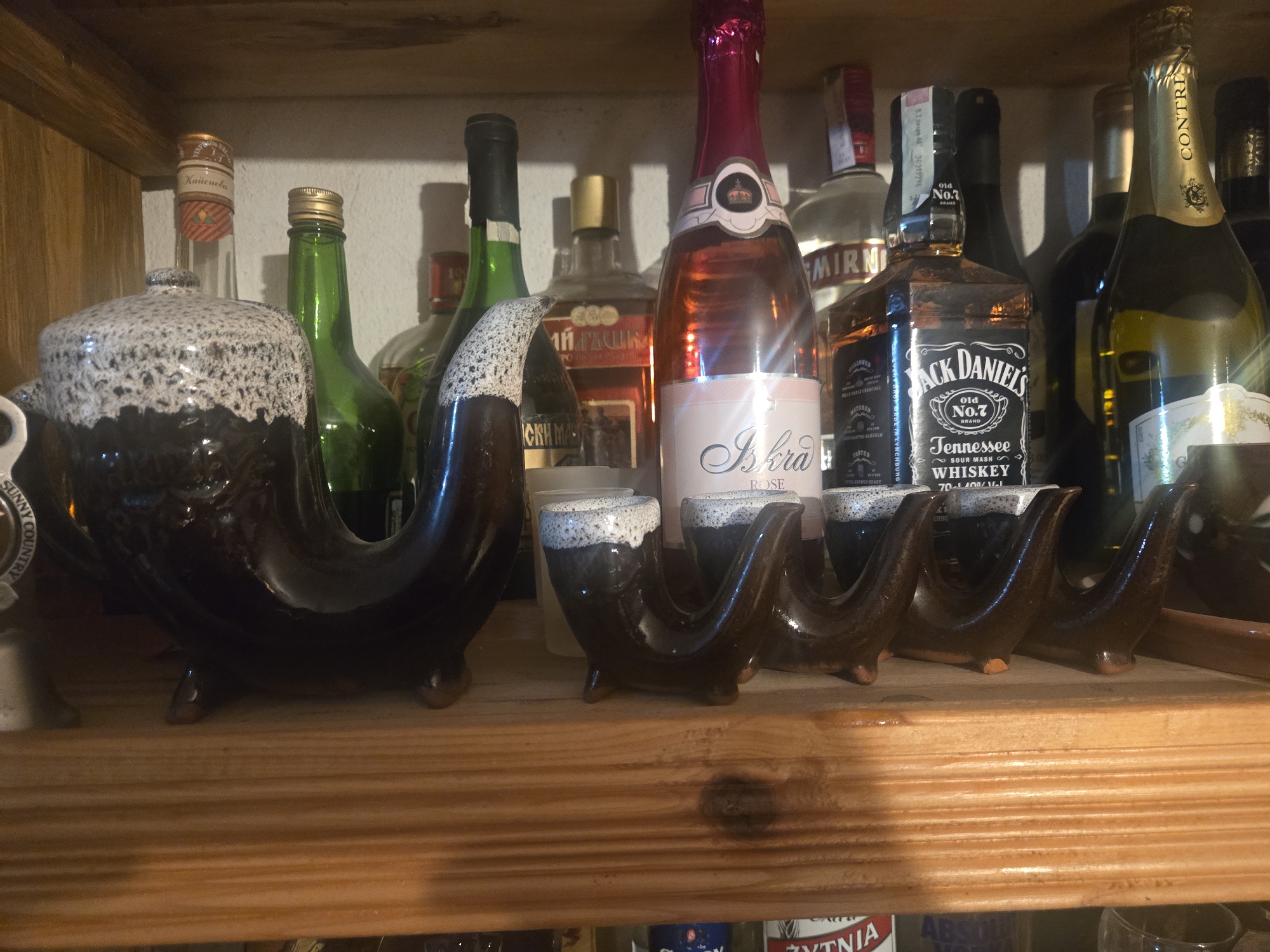 A shelf contains bottles of alcohol and a ceramic decanter and shot glasses. The bottles include "CONTRI", "Jack Daniel's Tennessee Whiskey", "Iskra ROSE", "МИ ГЪШКА", and "Кайсиева". The shelf is made of stained wood.