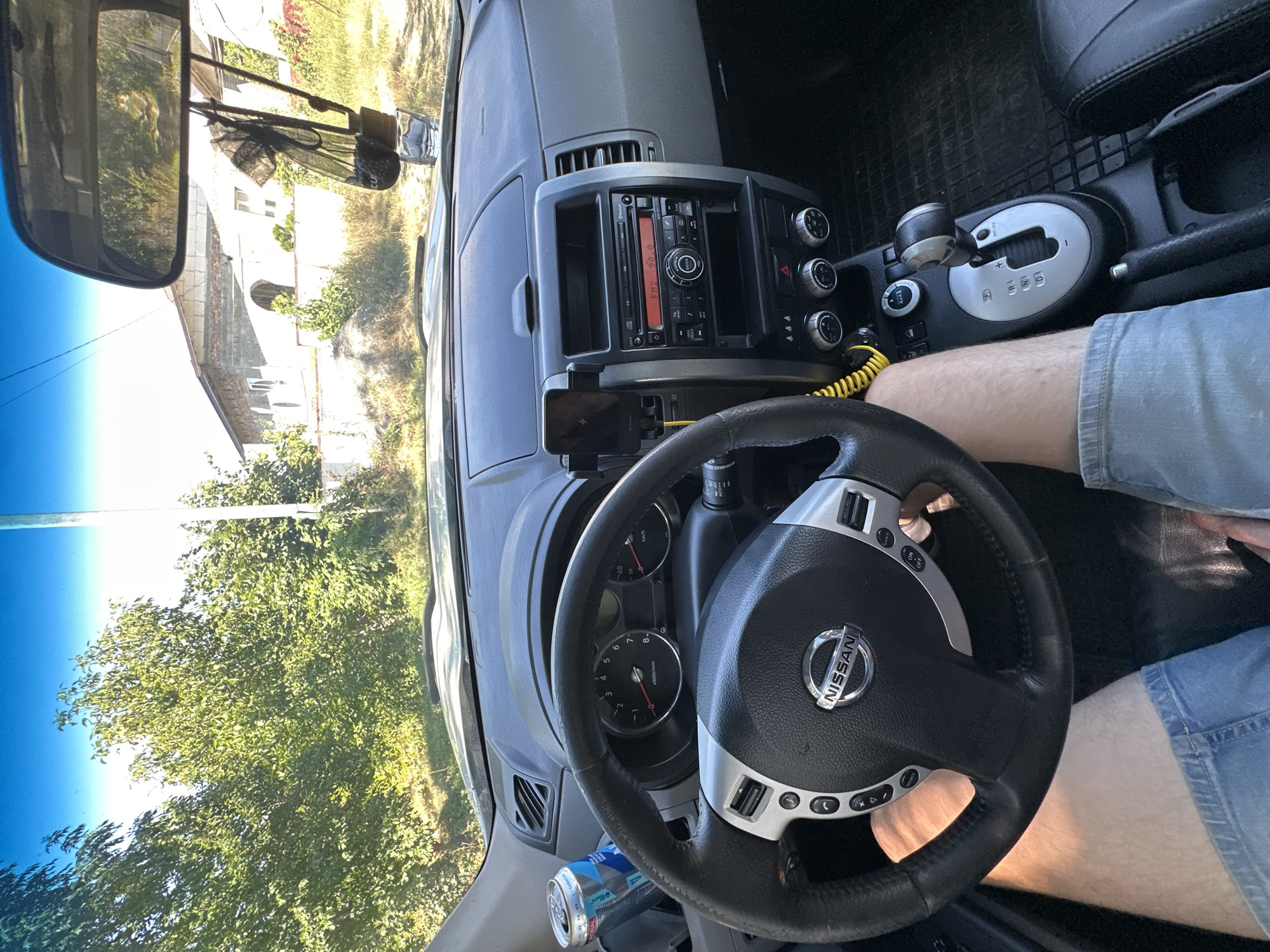 The image shows the interior of a Nissan car, taken from the driver's perspective. The steering wheel is visible in the foreground, with the Nissan logo in the center. The dashboard features a car radio, air vents, and various controls. A can of beverage is placed in a cup holder to the left of the steering wheel. The gear shifter is visible to the right, in the 'P' position. The driver's legs, clad in denim shorts, are visible. Outside the car, through the windshield and side windows, a tree-lined street can be seen, with houses in the background. The scene is set during the daytime, under a clear blue sky. The location is identified as Makiv, Ukraine.