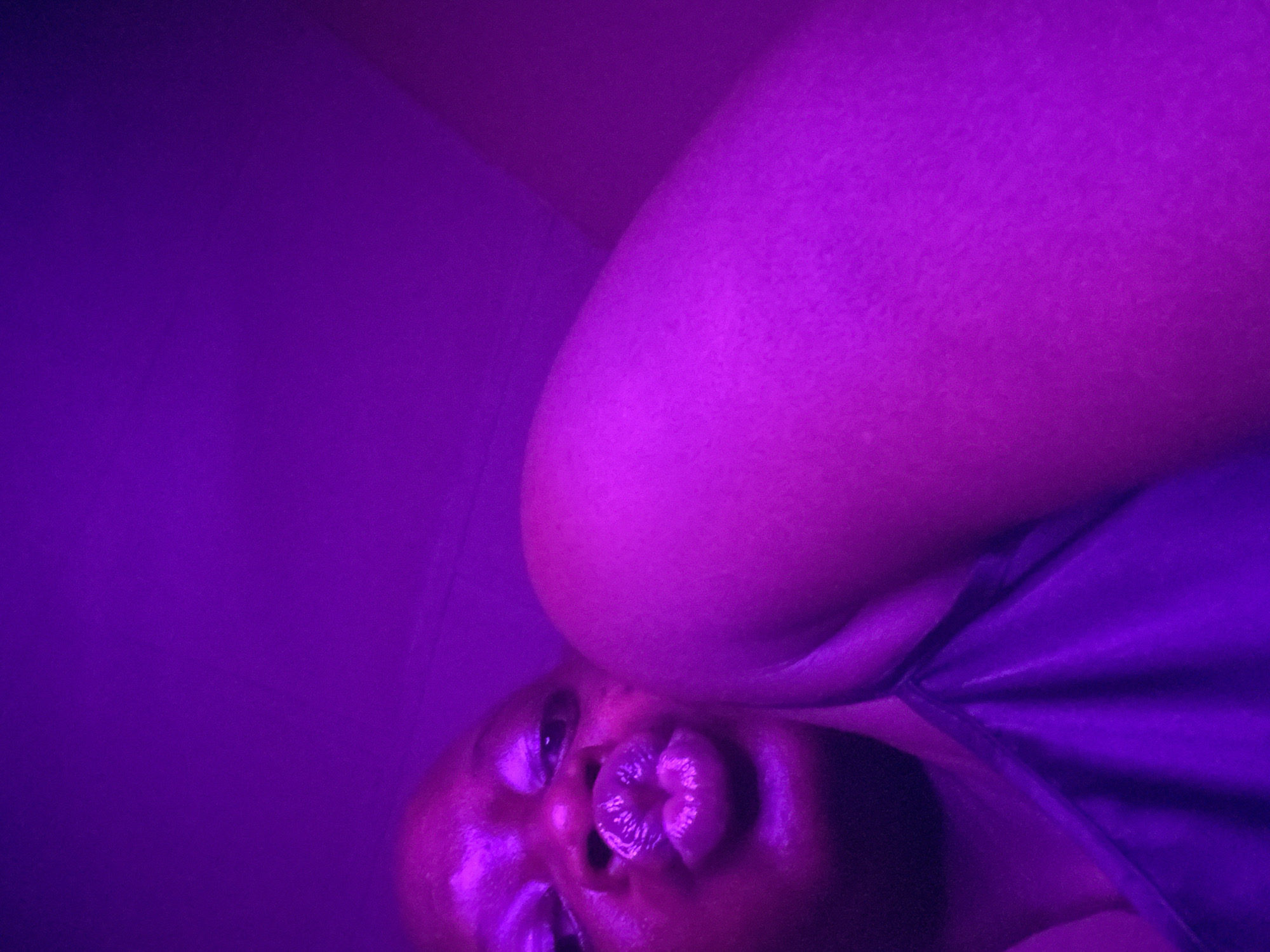 The image captures a close-up portrait of a person in an environment dominated by intense purple lighting, presumably in Maigizo, Nigeria. The subject, who appears to be a person with dark skin, is positioned at an angle, looking up towards the camera. Their face occupies the lower right portion of the frame.
Notable details of the subject include their dark eyes, which are open and directed at the viewer. Their lips are distinctively pursed in a "kissy face" or pouting expression, which is highlighted by the purple light, creating a glossy appearance on the surface of the lips. A portion of their upper body, specifically a shoulder or upper arm, is visible in the upper right, partially obscured by the angle and lighting. The fabric of what appears to be clothing, possibly a top or dress, is also visible around the subject's neck and shoulder area, reflecting the purple hue.
The setting is minimalist, with the background consisting of a plain, uniform vibrant purple color, suggesting a wall or simple backdrop. The pervasive purple light lends an artistic, moody, or club-like ambiance to the scene. The activity taking place is a posed photograph, with the subject intentionally making a facial expression. There are no visible objects other than the person and their attire, and no visible text anywhere in the image. The time of day or specific weather conditions are not discernible due to the artificial lighting.