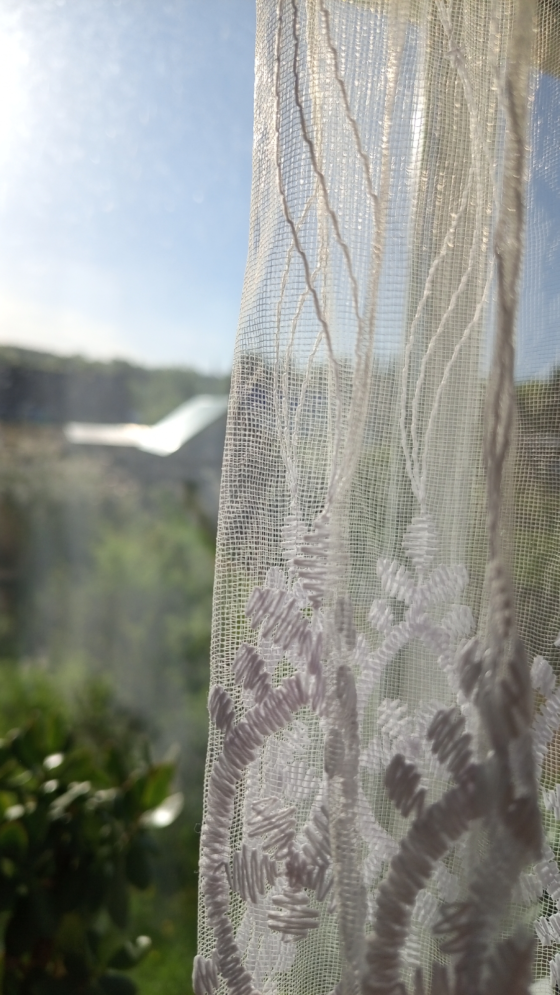 A close-up shot shows a white lace curtain partially obscuring a view of the outdoors. The curtain features a patterned design of embroidered shapes. Behind the curtain, the landscape is blurred, showing a green, slightly hilly terrain and a few buildings in the distance, under a bright, cloudy sky. There is no indication of the image being taken in the city of Уральское, Russia.