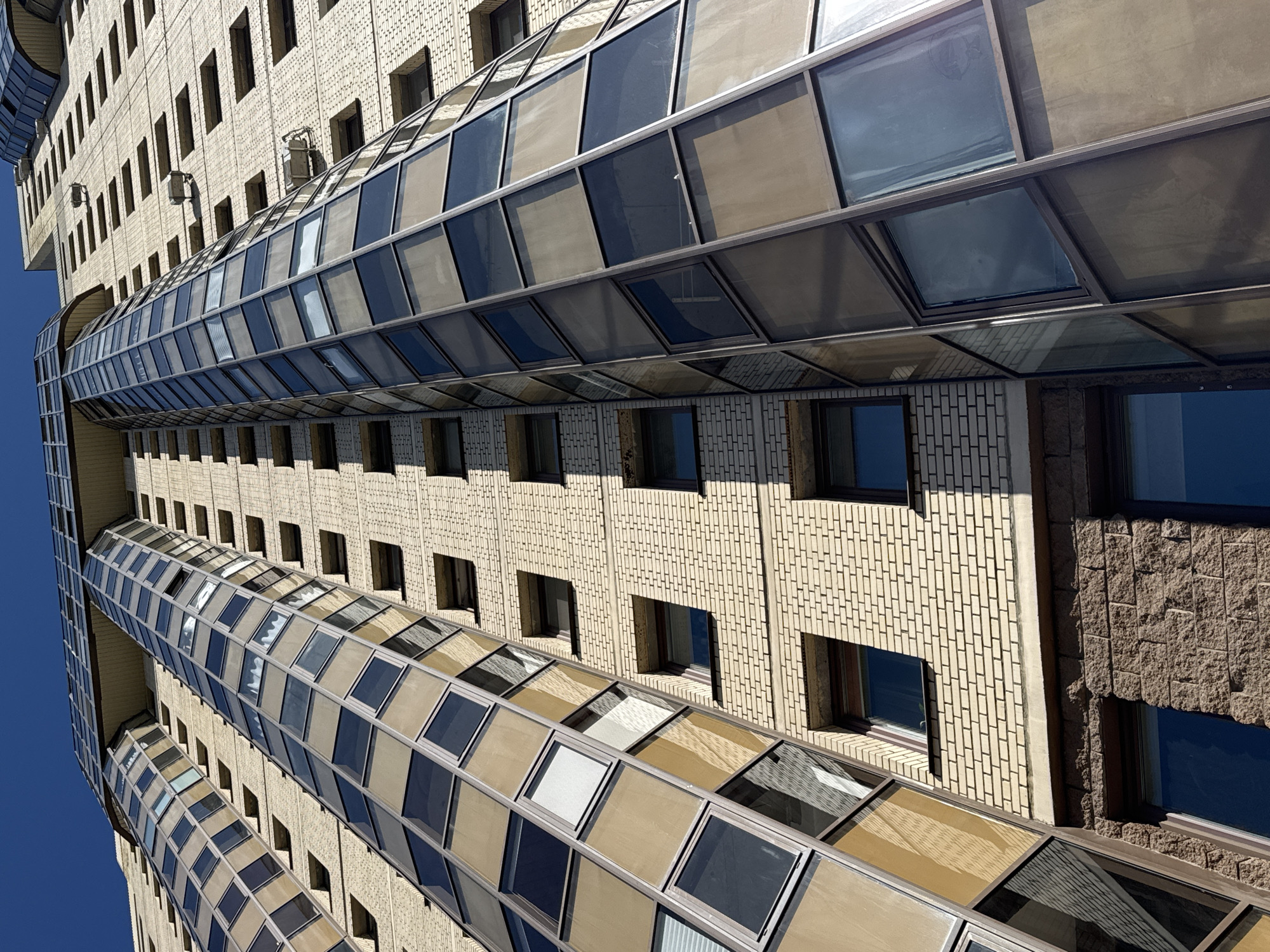 The image captures a view of a curved, multi-story building in Vsevolozhsk, Russia. The building features a mix of brick and stone textures, with numerous rectangular windows. A glass-enclosed walkway extends between sections of the building. The sky is a clear blue.