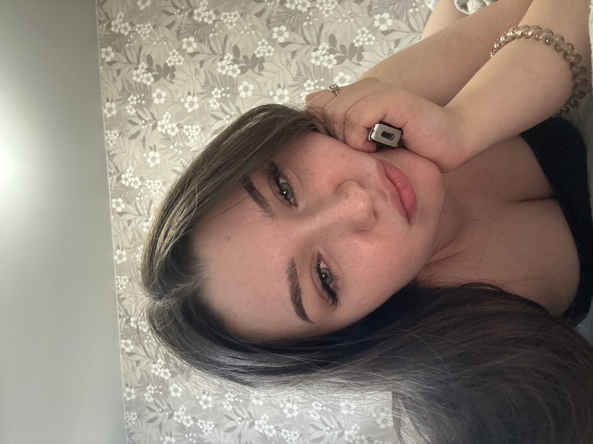 The image is a close-up, sideways selfie of a young woman lying down. She has long, dark brown hair and is wearing a black top. Her left hand is resting on her face, near her eye, and she is wearing a ring on her index finger. On her right wrist, she has a beaded bracelet. The background is a light-colored wallpaper with a floral pattern. The lighting suggests it is daytime, possibly indoors, given the wallpaper. Her expression is relaxed and serene. There is no visible text in the image. The location context is Zarechnaya (Gamovskoe S/P), Russia.