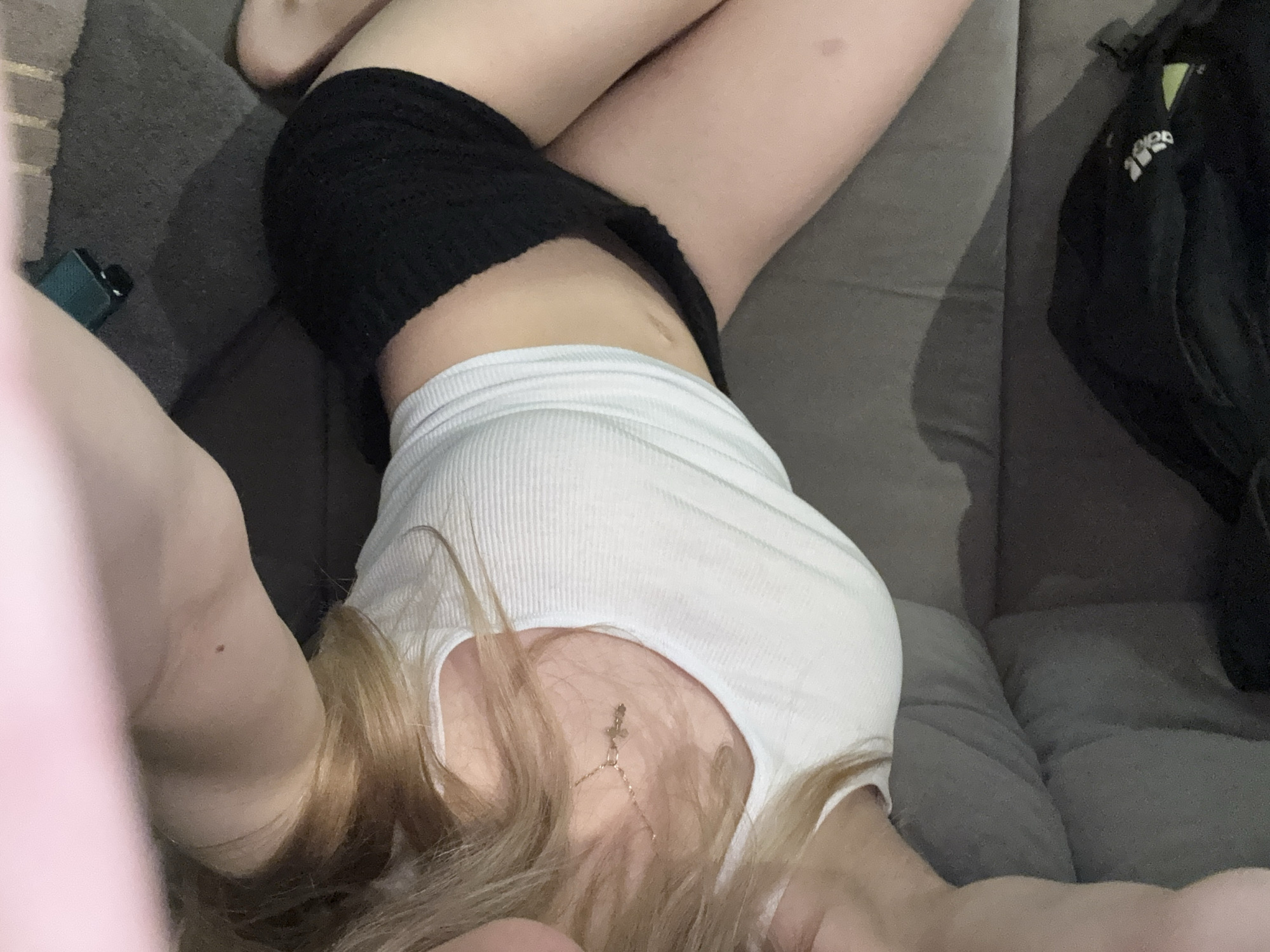 A person is lying on a grey sofa. The person is wearing a white tank top and black shorts. A black Adidas bag is on the right side of the sofa. The location is unknown.