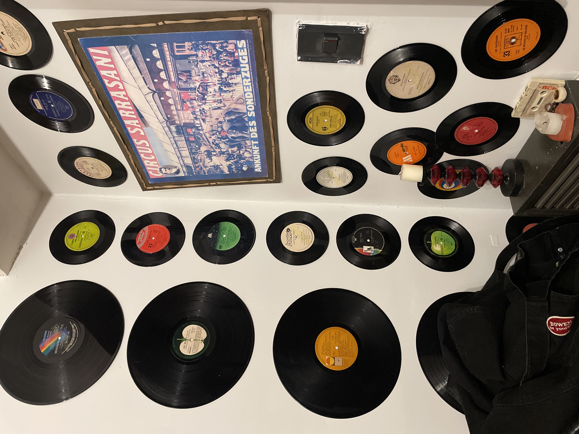 The image presents a collection of vintage vinyl records and related memorabilia arranged on a white wall. The records vary in size, from smaller 45s to larger LPs. Labels visible on some of the records indicate artists, song titles ("Amarillo," "Los Naufragos," "De Boliche en Boliche"), and record labels (CBS, RCA, Micsa). Interspersed among the records are additional items: a framed circus poster labeled "Circus Sarrasani" and "Ankunft des Sonderzuges," a black electrical outlet, a vintage RCA cassette tape, a small candle holder, and a decorative glass/crystal ornament. A black denim jacket with a "Bower be your" patch is partially visible on the right side of the image, obscuring some of the records. The setting appears to be indoors, but the location in the city of Buenos Aires, Argentina, cannot be determined from the image alone.