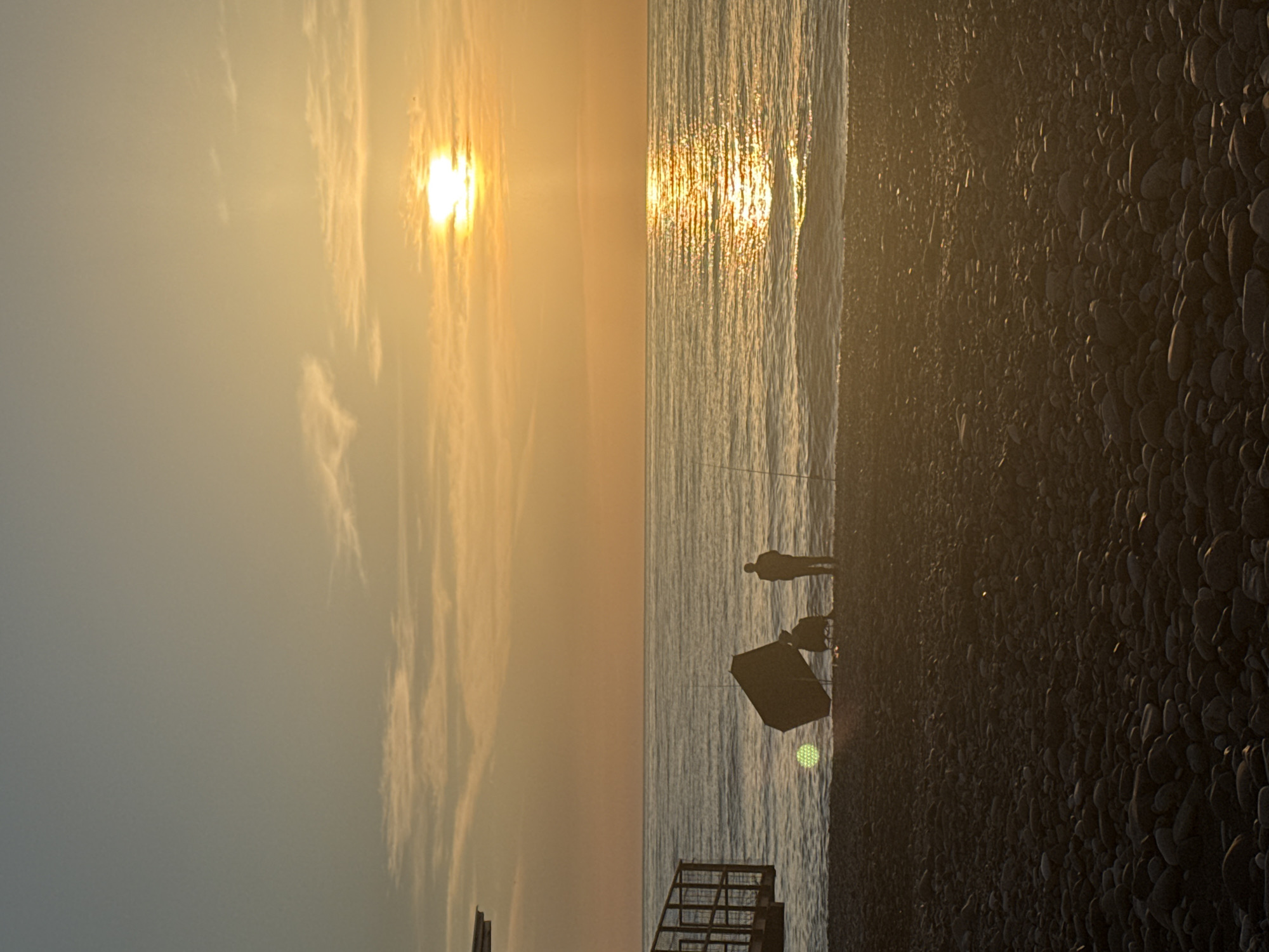 The image depicts a sunset scene at the beach in Batumi, Georgia. The sky is a gradient of light blue to orange, with clouds. The sun is setting over the Black Sea, and its reflection can be seen on the water. A man is standing near the water's edge, fishing. A beach tent is next to him. In the foreground, there are pebbles, and what appears to be a metal staircase is visible at the bottom. The image has a warm and calm atmosphere.