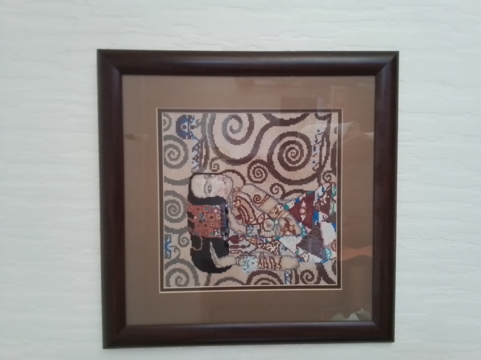 A framed cross-stitch depiction of Gustav Klimt's "The Kiss" hangs on a textured, off-white wall. The artwork is housed in a dark brown wooden frame with a beige mat. The cross-stitch pattern captures the iconic embrace of the couple, with intricate details in their clothing and the swirling background. The lighting suggests an indoor environment.