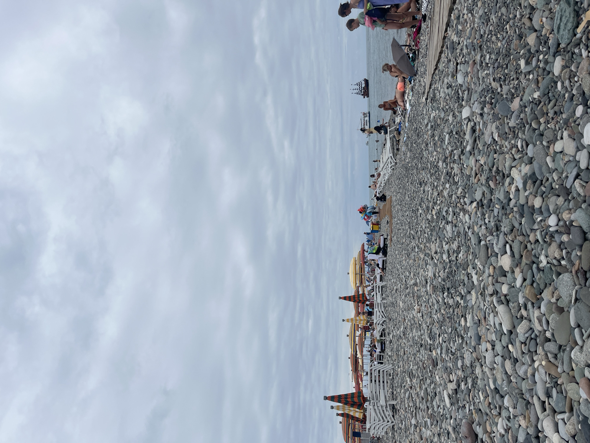 The image depicts a crowded beach scene with a prominent foreground of grey pebbles. In the middle ground, rows of white lounge chairs and colorful striped umbrellas are set up, some occupied by sunbathers. Several people are visible, some reclining on chairs, others walking or interacting. The background shows the sea under an overcast sky, with a few boats on the water, including what appears to be a pirate-style ship. The overall impression is of a busy, somewhat overcast day at a pebble beach, likely in Batumi, Georgia, a popular seaside resort known for its beaches and tourist attractions.