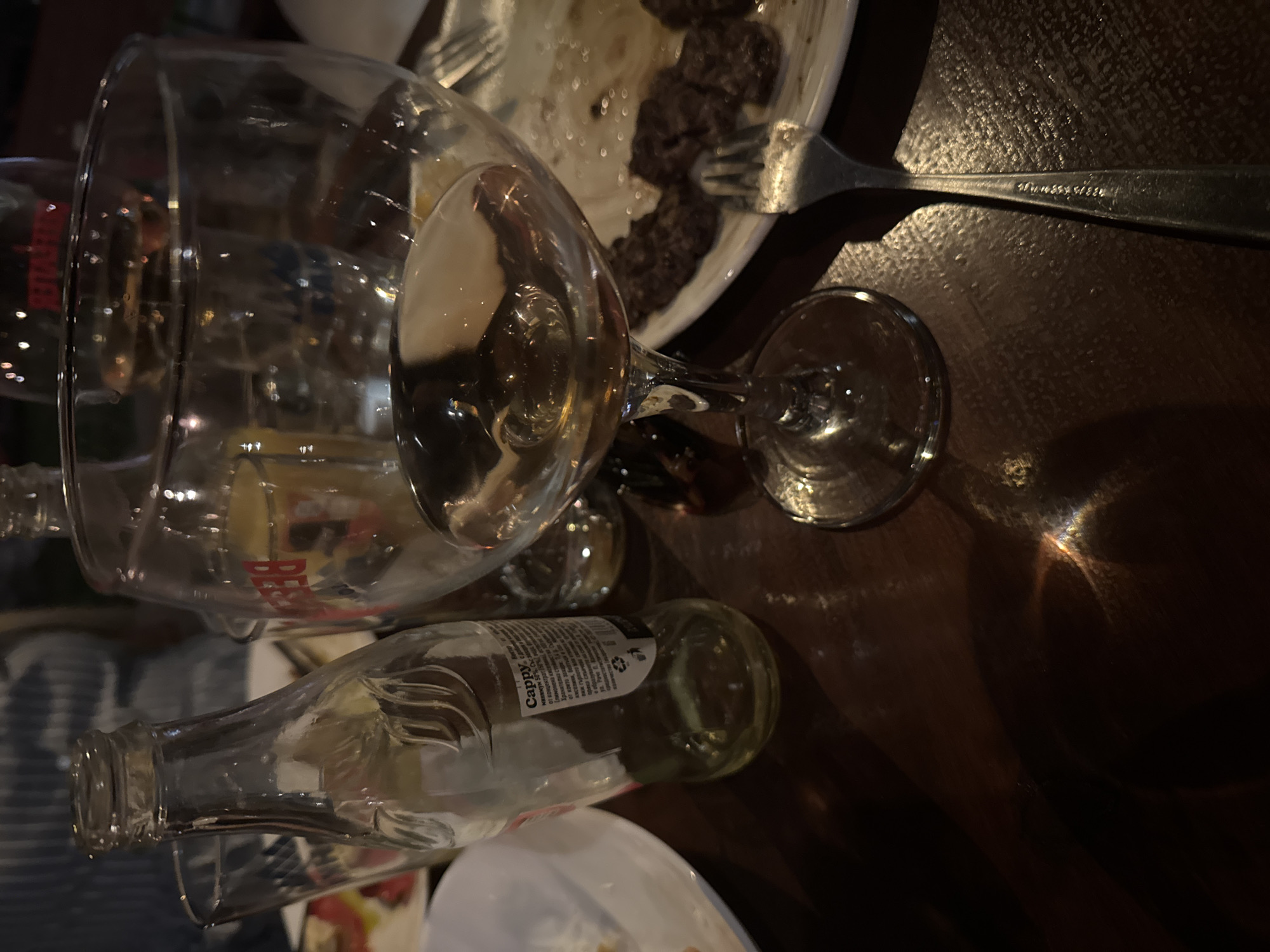 A still life image captures a table setting with several beverages and food remnants. In the foreground, a clear wine glass contains a pale yellow liquid, possibly white wine or a similar beverage. To its left, a small, clear glass bottle, partially filled with the same liquid, stands upright. Its label, partially visible, suggests the brand "Cappy." Behind these, another glass with a red logo is visible, and a third glass with a blurred blue design can be seen further back. A white plate holds several pieces of cooked meat, appearing to be grilled or pan-fried, along with what looks like a fork. The lighting in the scene is dim, with light reflecting off the glassware, creating streaks of color on the dark wooden tabletop. The overall impression is of a meal that has recently concluded or is in progress, set in a dimly lit indoor environment. The location in Dimitrovgrad, Bulgaria, is inferred from the Cyrillic text on the bottle label.