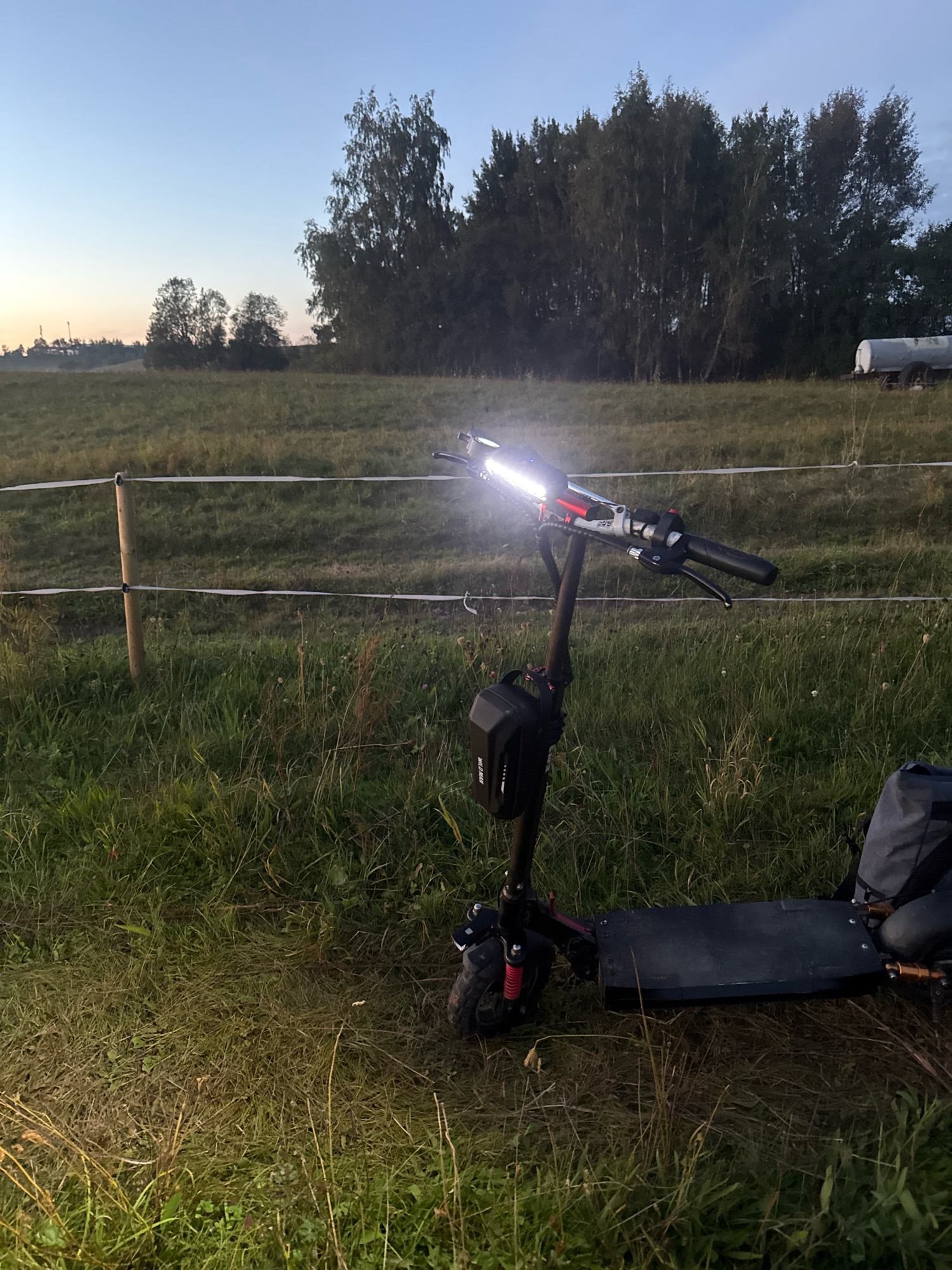 An electric scooter with its headlight on is parked in a grassy field at dusk. The scooter is dark-colored with red accents and has a small black bag attached to its stem. In the background, there is a line of trees and a water tank on wheels. The sky is a pale blue, indicating the end of the day. The location is the village of Galinė, Lithuania.