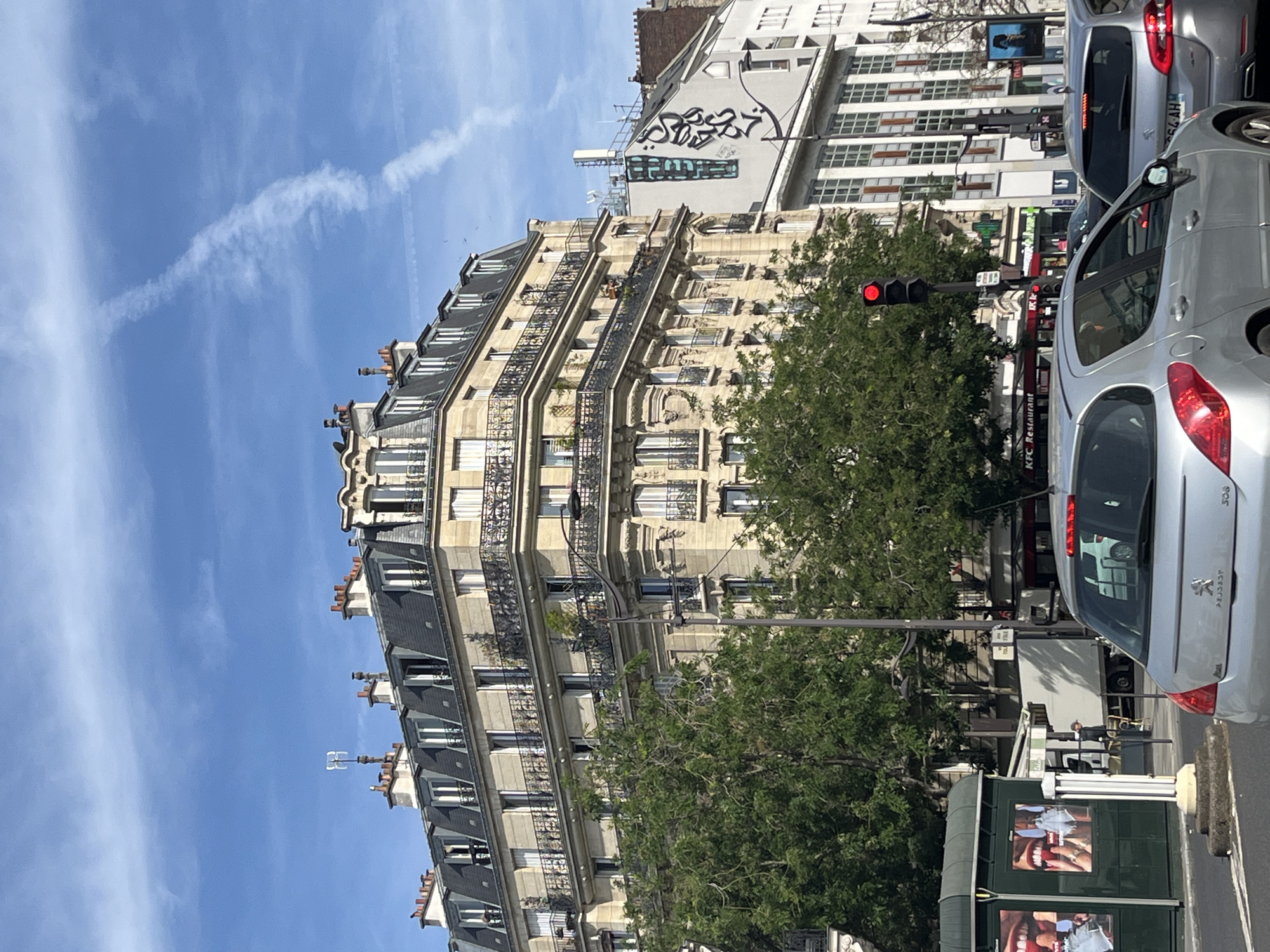 This image captures a street scene in Paris, France, likely during the day. The foreground features the rear of a silver Peugeot 308 hatchback, followed by another silver hatchback, and a white truck in the background. A green bus shelter with advertisements is visible on the left.
Dominating the midground and background is a large, ornate Haussmann-style building with a mansard roof, typical of Parisian architecture. This building has multiple stories, balconies with wrought-iron railings, and numerous windows. Trees with lush green foliage partially obscure the lower floors of the building. To the right of this building, a more modern, white building stands, featuring a grid-like facade and graffiti on its upper level.
A traffic light shows a red signal. Various signs are visible, including one for "KFC Restaurant." The sky is blue with scattered white clouds and a prominent contrail. The overall impression is of a busy urban environment in Paris.