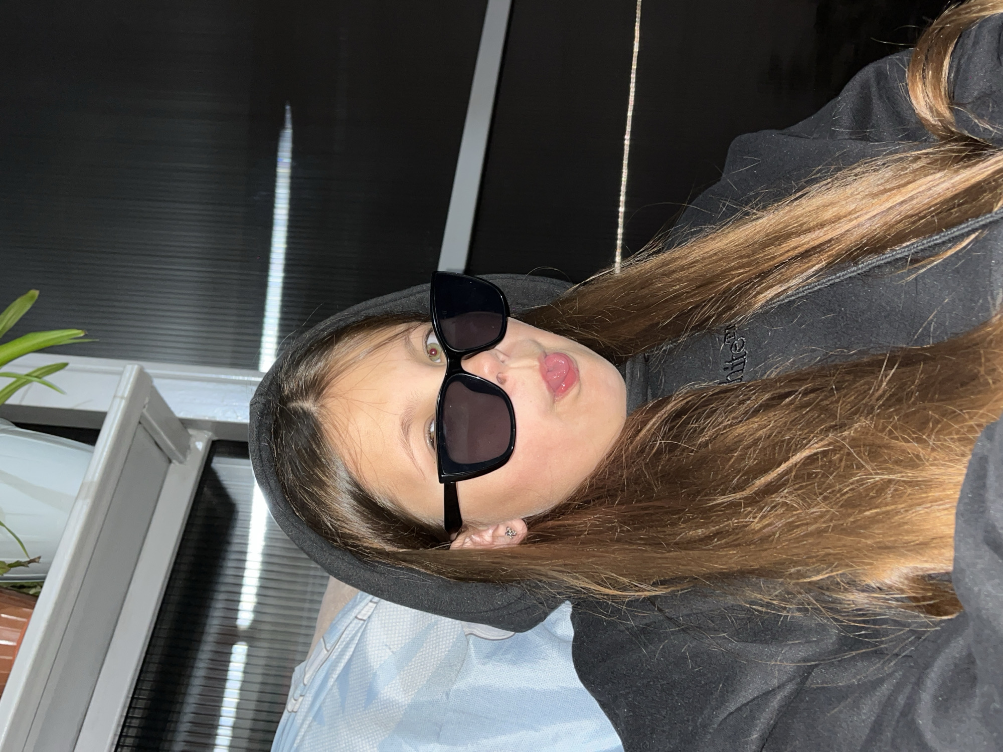 A girl with long brown hair, wearing a black hoodie and black sunglasses, is sticking her tongue out. An earring is visible in her left ear. The background features a plant, a window, and possibly a metal blind. The image appears to be taken indoors. There is no information in the image to determine the location in the city of Novopokrovskaya, Russia.
