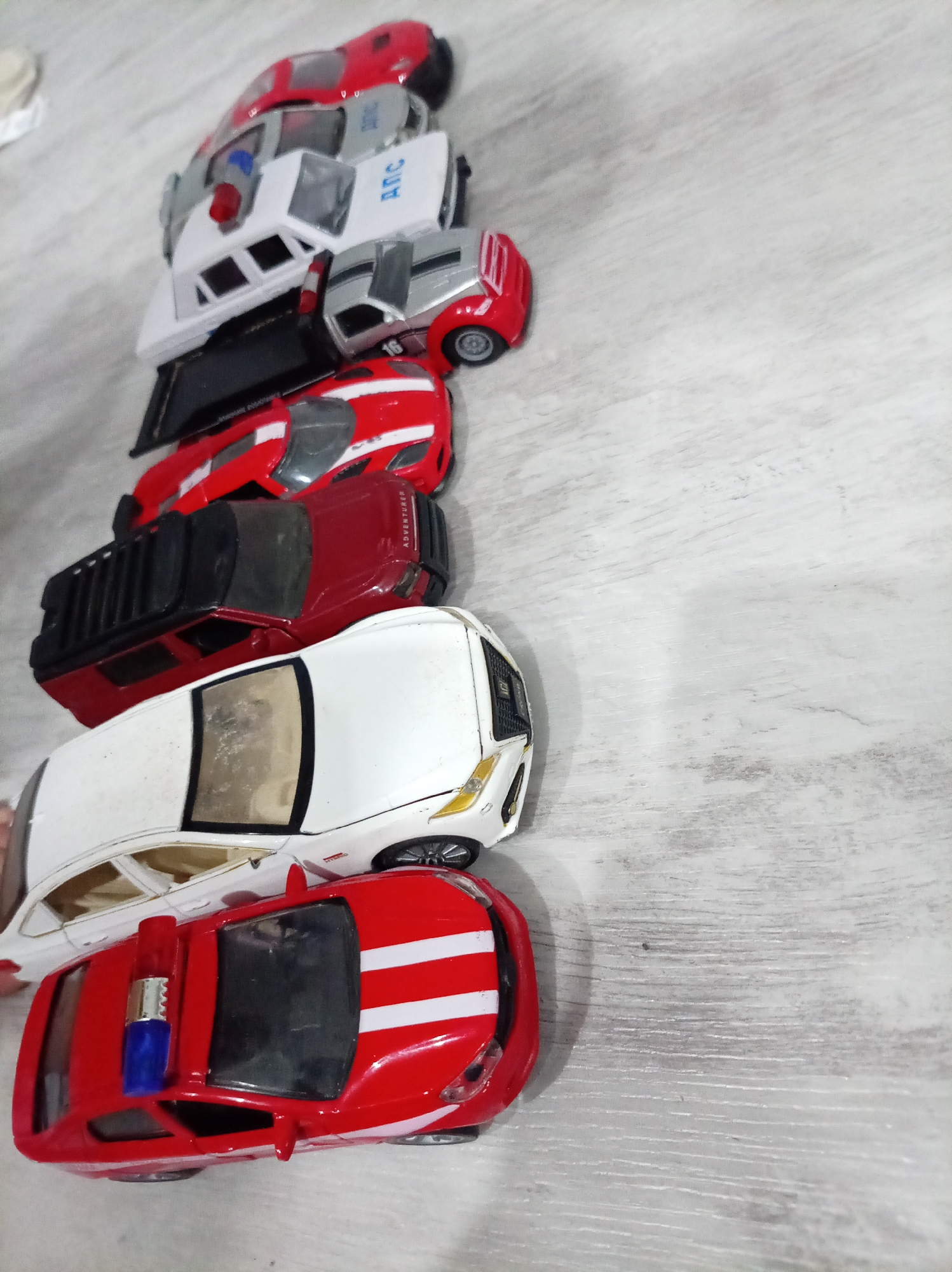 A line of toy cars is displayed on a light-colored, wood-grain textured surface. The cars are arranged diagonally from the bottom left to the top right of the frame. Starting from the bottom, there is a red toy car with white stripes and a blue and red flashing light on top. Next is a white toy car with black tinted windows and gold accents on the front. Above that is a red toy SUV with a black rack on top and the word "ADVENTURER" printed on the side. Following this is a red toy car with white stripes. Then, a silver toy car with red accents. Above that is a white toy police car with red and blue lights and the letters "ANC" on the side. The topmost toy car is a red one with a silver accent on the rear. The overall impression is a collection of toy vehicles. The image does not contain any elements that suggest a specific location within Oktyabrsk, Russia.