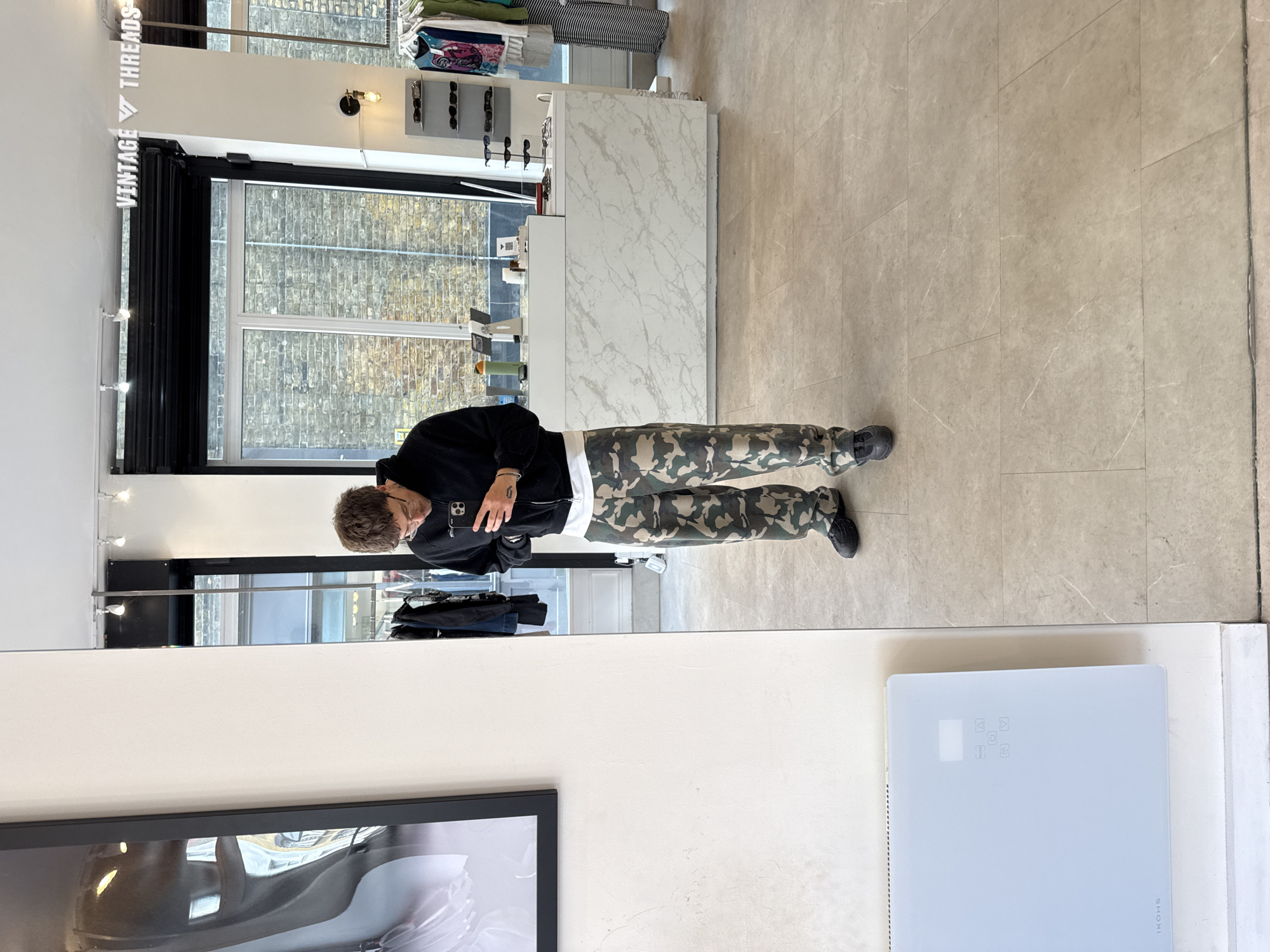 A person stands in a retail setting, likely within the city of London, United Kingdom, based on the "Vintage Threads" sign visible at the top left. The person wears a black jacket, a white shirt or under-layer, and camouflage pants, facing away from the camera and towards a mirror. The reflection in the mirror shows the person holding a phone, potentially taking a photo of themselves. The setting appears to be a shop with a window to the left revealing brick buildings across the street. To the rear of the person and within the store, there is a countertop display, which contains various items. A framed image is visible in the lower left, and a white appliance is in the lower right, marked with "IKOHS". The floor is tiled.