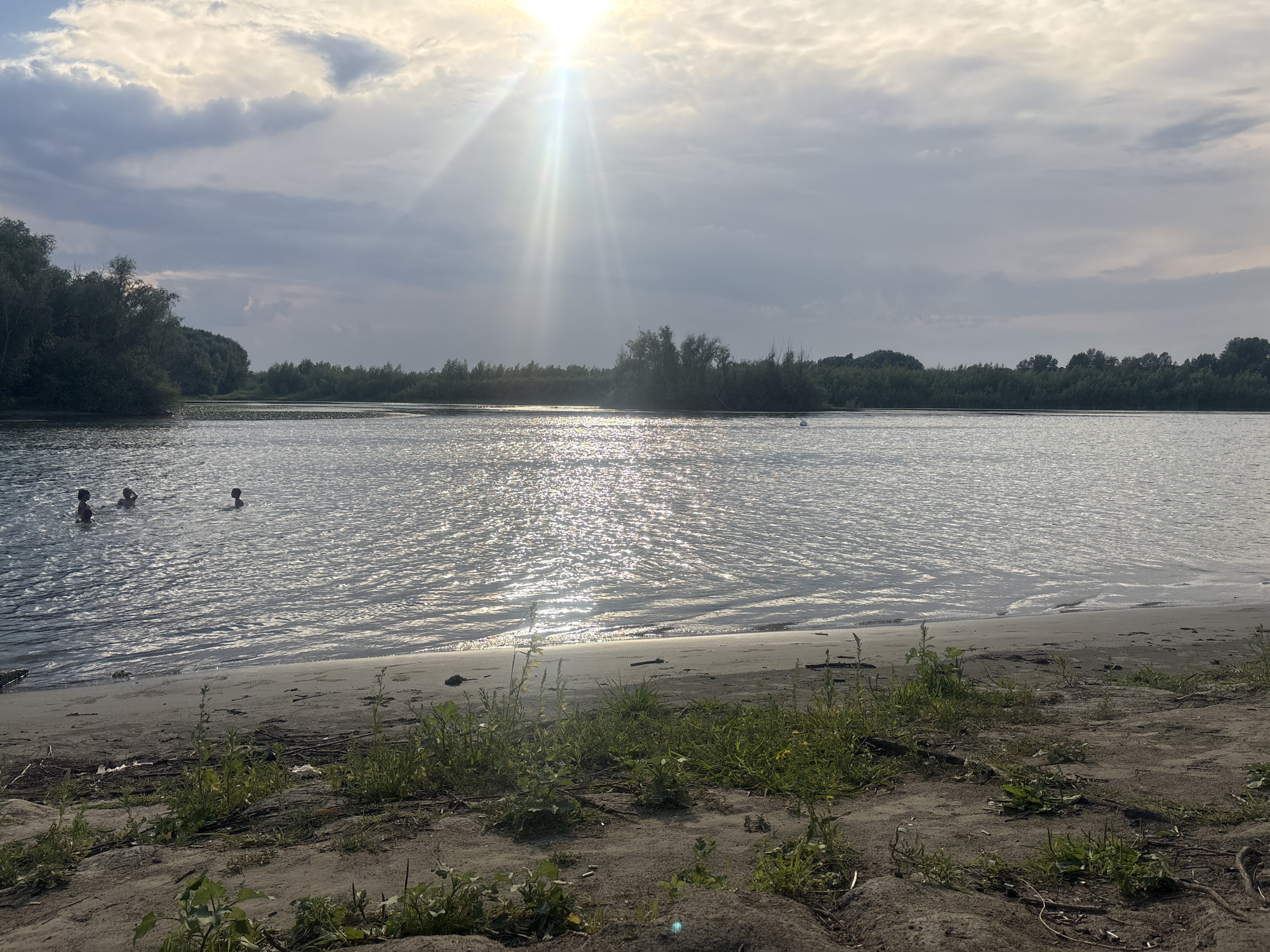 This image depicts a wide, calm river with a sandy bank in the foreground. The sun is shining brightly through scattered clouds, casting a shimmering reflection on the water's surface. Several people are visible in the river, appearing to be swimming or bathing. The far bank is lined with dense green trees, suggesting a natural or semi-natural environment. The overall scene conveys a peaceful and recreational atmosphere, possibly during a warm day. This location is likely a river beach or a natural swimming area within or near the city of Pavlodar, Kazakhstan, given the presence of a large river and the general landscape.