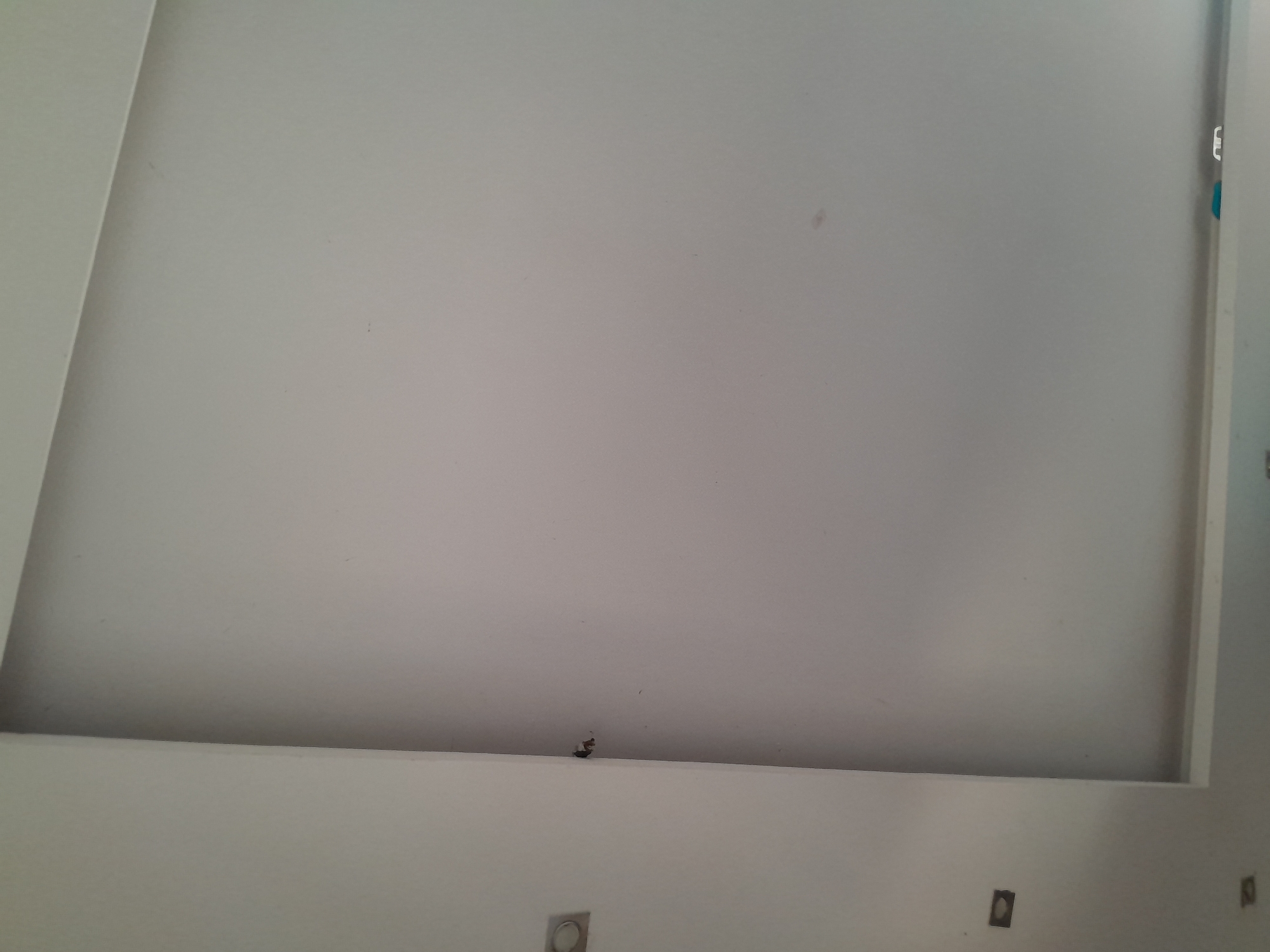 The image shows a close-up view of a white ceiling with a recessed section. On the left side of the image, there are two square recessed fixtures, possibly for lights, mounted on a white wall that meets the ceiling. In the corner where the wall meets the ceiling, there is a small dark mark or stain. The recessed section of the ceiling appears to be a darker shade of white than the main ceiling area, creating a shadow or depth effect. Towards the bottom of the image, along the edge of the recessed ceiling, there are two small, colorful objects, one blue and one white, which appear to be some form of plastic fixture or cap. The overall scene suggests an interior architectural detail. The location is identified as Santa Ana, Venezuela.