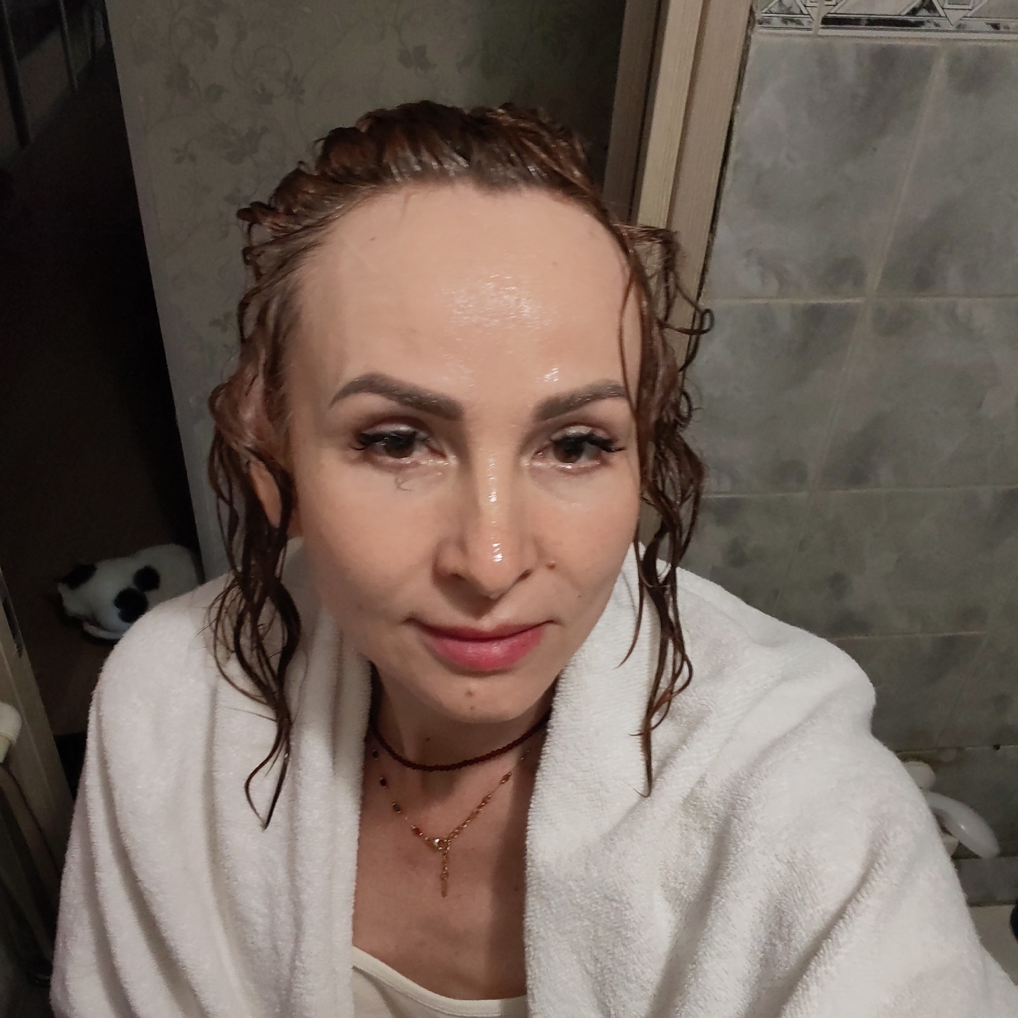 A woman with wet auburn hair, wearing a white towel, is posing indoors. She has brown eyes and fair skin. She is also wearing two necklaces.
Behind her is a wall with light-colored floral wallpaper to the left, and a wall tiled with gray square tiles to the right. In the lower left corner of the shot is a black and white object, possibly a stuffed animal or pet, sitting on the floor. The location is consistent with an interior in the city of Chaikovsky, Russia.
