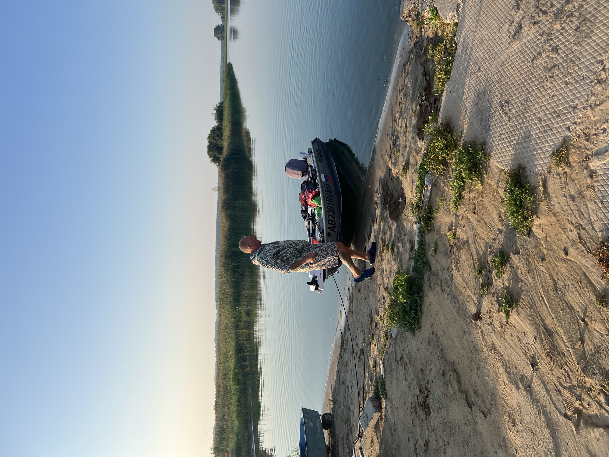 A bald, fair-skinned man wearing a camouflage hoodie and shorts walks along a sandy bank toward a small motorboat. The boat is equipped with a trolling motor and has several bags and red containers loaded into it. The man is walking in shallow water, and his legs are wet. The boat is tied to the shore with a rope attached to a concrete block. Another small boat on wheels is partially visible on the left side of the image. The scene takes place on a clear, sunny day by a calm body of water, likely a river or lake, with a lush green shoreline in the background. The location is Zav'yalovo, Russia.