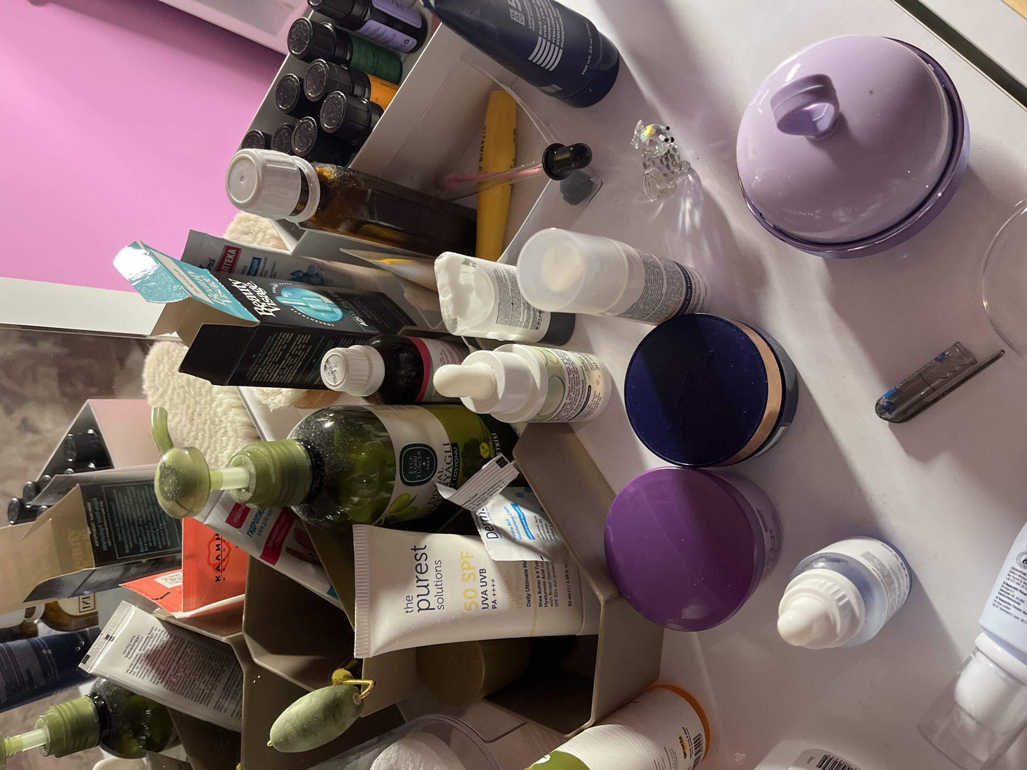 The image shows a close-up, disorganized collection of personal care and beauty products. Items include skincare bottles with pump dispensers, tubes of sunscreen and moisturizer (one labeled "the purest solutions 50 SPF UVA UVB"), dropper bottles, and cosmetic compacts. There is an opened box of "Beauty Visage AQUA-FILLER." Several bottles are of brown glass. The products are scattered across a white surface. The text on a green glass bottle indicates it contains products of "EYÜP TUNCI" from the year 1923. There is no information to suggest this is from Alanya, Turkey. There are no people present in the image.