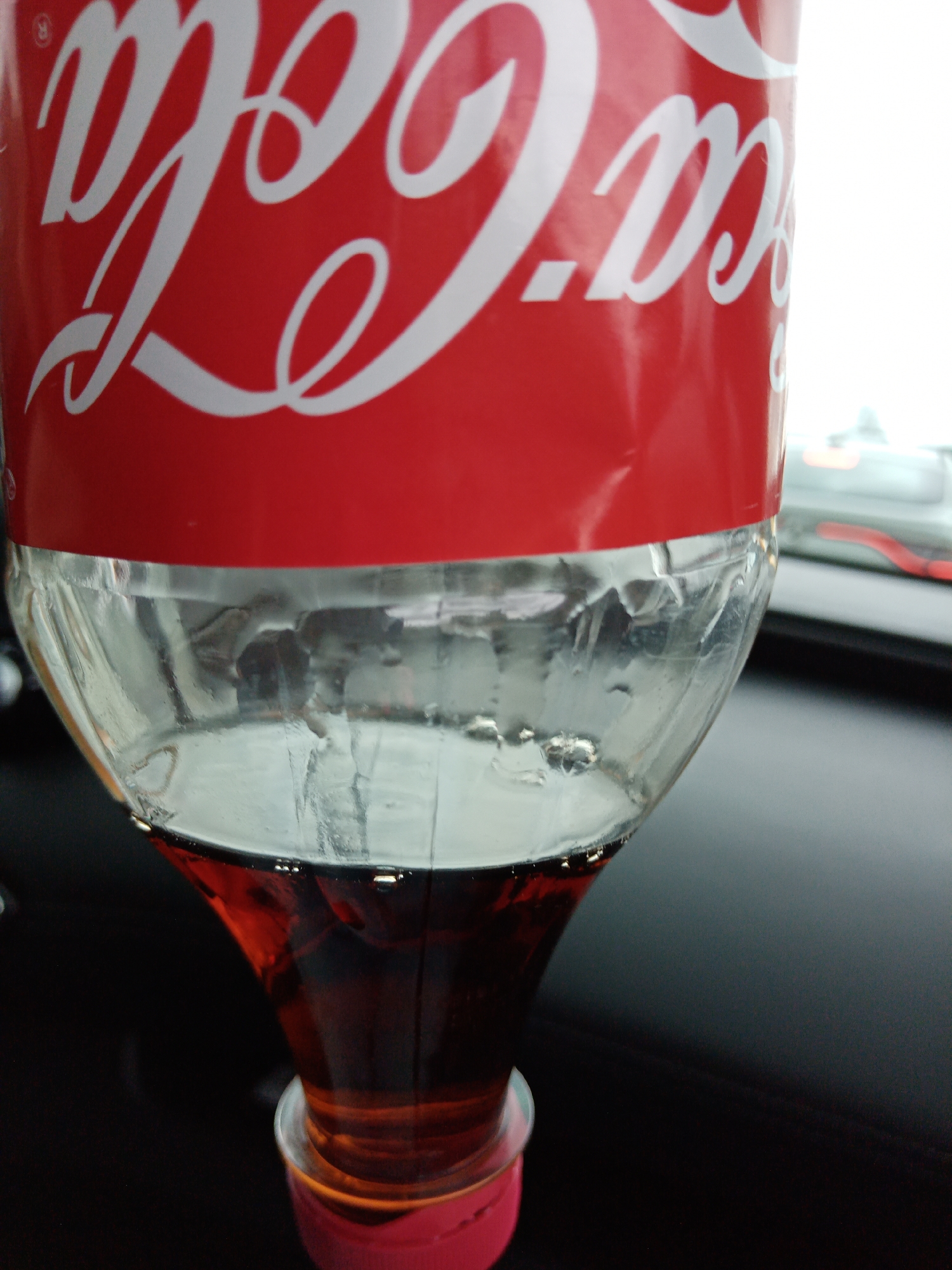 The image shows a partially consumed bottle of Coca-Cola. The top portion of the bottle displays the Coca-Cola logo in white letters against a red background. The lower portion of the bottle is clear plastic and contains the remaining dark-colored soda. The bottle appears to be sitting inside a vehicle, with a dark dashboard visible in the background. No people are visible in the frame. There is no evidence to indicate the location is Kriulino, Russia.