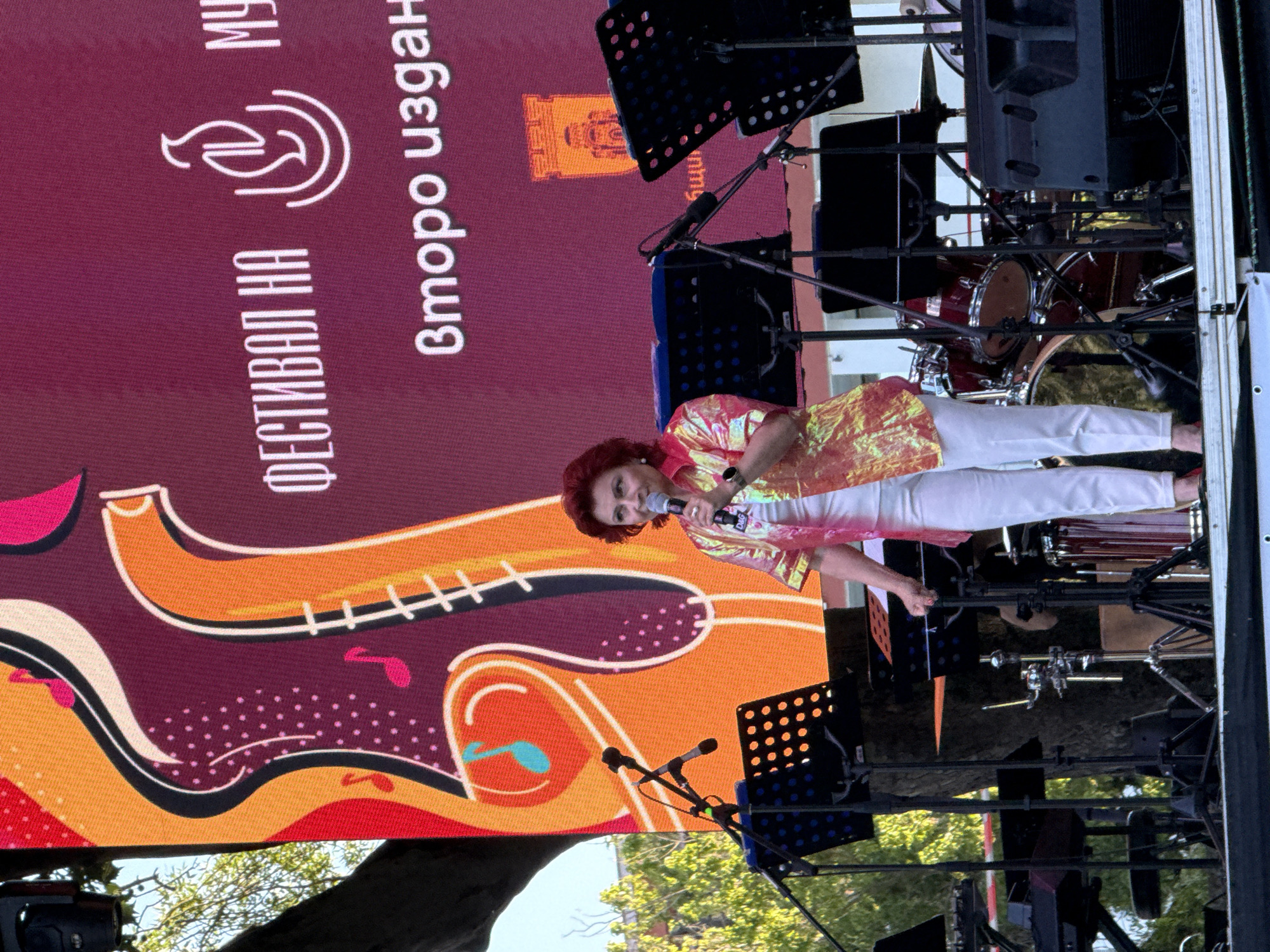 In Pleven, Bulgaria, a woman with short, red, curly hair is singing into a microphone on a stage. She is wearing a shiny, iridescent orange and yellow patterned shirt over a white top and white pants. Behind her, a large LED screen displays a vibrant, colorful abstract design with musical notes and saxophone-like shapes. Text on the screen, in white, reads "ФЕСТИВАЛ НА МУ" and "Второ издан". A logo resembling a stylized flame or hand is visible on the screen. The stage is equipped with several microphones on stands, music stands, and musical instruments, including a drum set. The lighting suggests it is daytime, with sunlight filtering through trees in the background. The overall scene depicts a live musical performance.