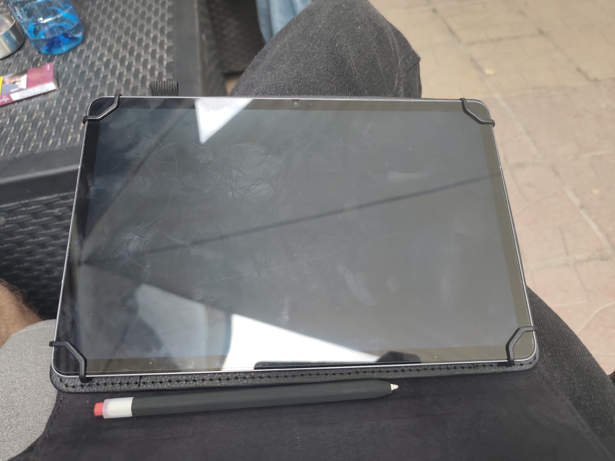 The image depicts a tablet resting horizontally on an individual's lap in the city of Lom, Bulgaria. The tablet, dark in color, is enclosed within a black, stitched folio case secured by four black corner elastic/rubber straps. Its screen is off or in standby mode, displaying significant smudges, fingerprints, and reflections of the surrounding environment. A small, circular front-facing camera is centered at the top bezel of the tablet. Below the screen, a dark gray stylus with a white tip and a red band is positioned on the tablet's case. Visible portions of the individual's bare left thigh and dark fabric (possibly denim) on the right thigh are present, indicating the tablet's placement on their lap. To the upper left of the tablet, on a dark, woven surface (likely outdoor furniture), a clear plastic bottle with blue liquid, a silver metal cup, and a red and white cigarette pack are visible. The background on the right side reveals a light brown or beige tiled ground surface.