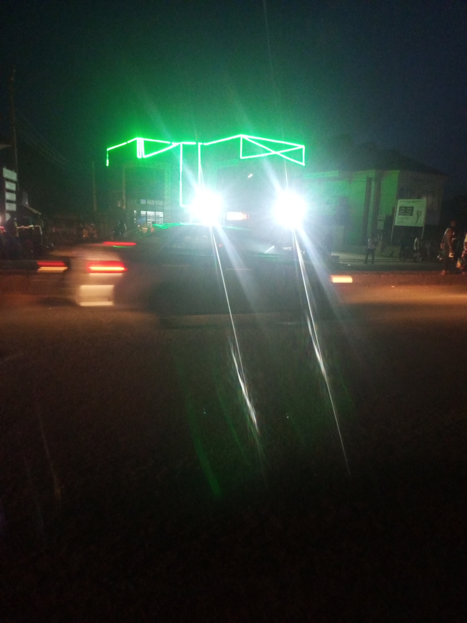 The image depicts a vibrant street scene at night in Sabon Tasha, Nigeria. In the immediate foreground, a vehicle, likely a car or small truck, faces the viewer with extremely bright headlights that create prominent streaks of light extending downwards onto the dark asphalt road. Dominating the upper part of this vehicle is an elaborate, geometric structure constructed from bright green neon or LED lights, casting a strong emerald glow onto the surrounding darkness.
Other vehicles are visible further down the road, indicated by blurred red taillights and hints of other lights, suggesting ongoing traffic. Pedestrians are scattered along the sidewalks on both sides of the street, appearing as blurry figures due to motion or low light conditions. Buildings line the street in the background, with some internal lights visible through windows, contributing to the urban night ambiance. Utility poles and wires can be vaguely discerned on the left.
The time of day is clearly night, with the artificial lights from vehicles and buildings serving as the primary illumination sources. The weather appears clear. The striking green light display on the central vehicle suggests it might be part of a local celebration, parade, or a specially decorated transport vehicle for an event in this busy street environment. A light-colored sign is visible on the right sidewalk near a building, but any text on it is unreadable due to distance and blur.