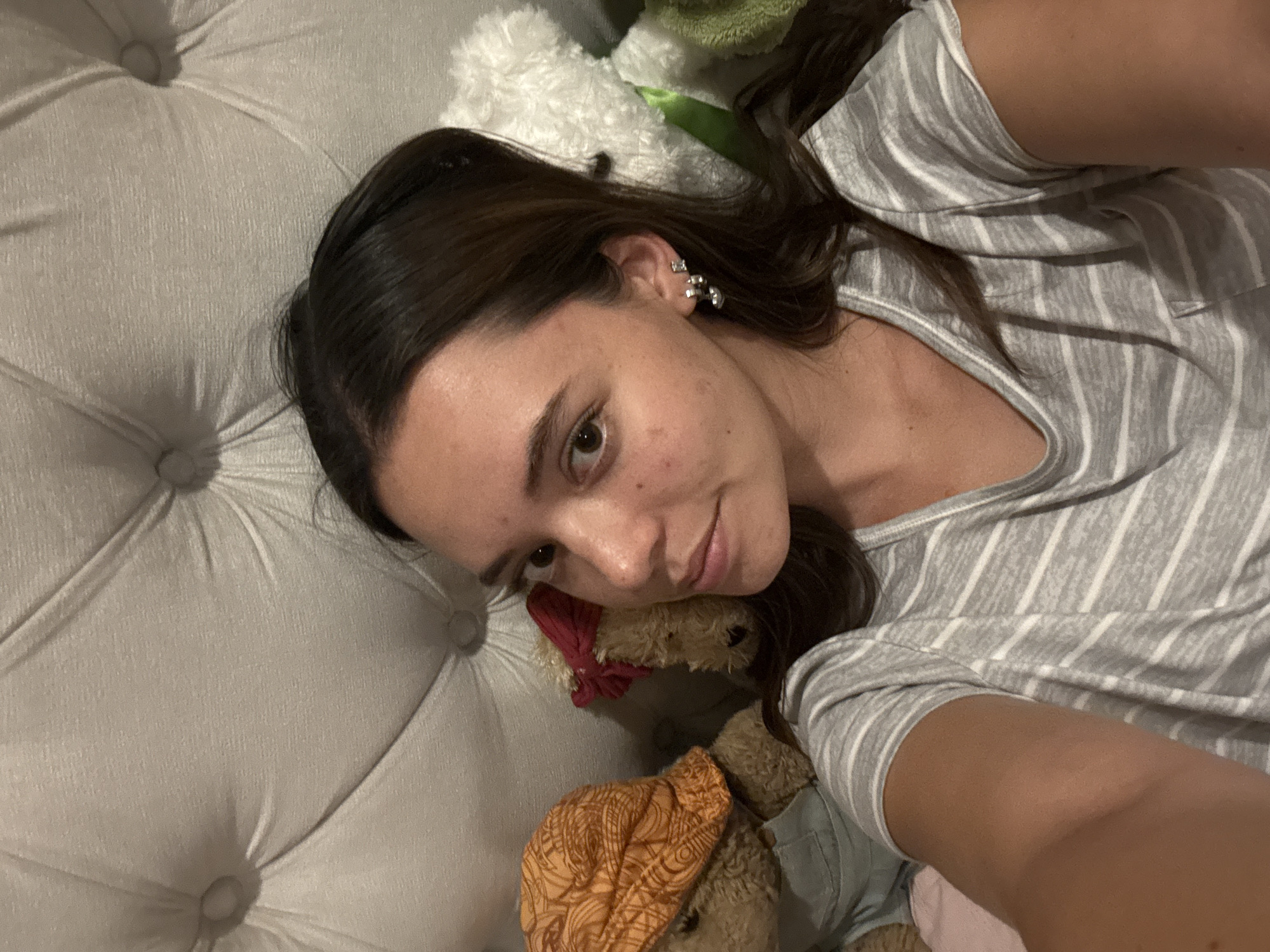 A woman is lying on a light gray, tufted cushion, possibly a headboard or sofa, with two teddy bears. She wears a gray and white striped top. One teddy bear has a red bow and the other has an orange patterned bandana. The woman has dark hair and earrings. The location in San Bernardino Tlaxcalancingo, Mexico cannot be determined from the image.