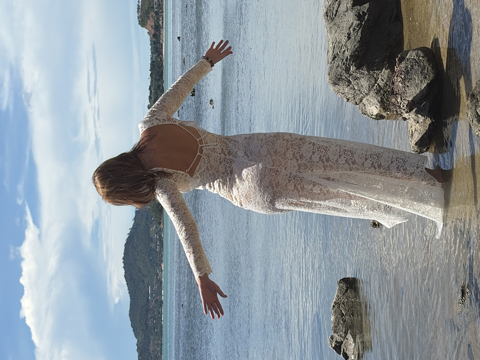 In Baan Bo Phut, Thailand, a woman stands in shallow ocean water near rocks on a beach. She is wearing a long-sleeved, white lace dress with an open back, adorned with pearls. Her arms are outstretched, and she is facing away from the camera, looking towards the sea and a distant, verdant island. The sky is partly cloudy, and the water is calm.