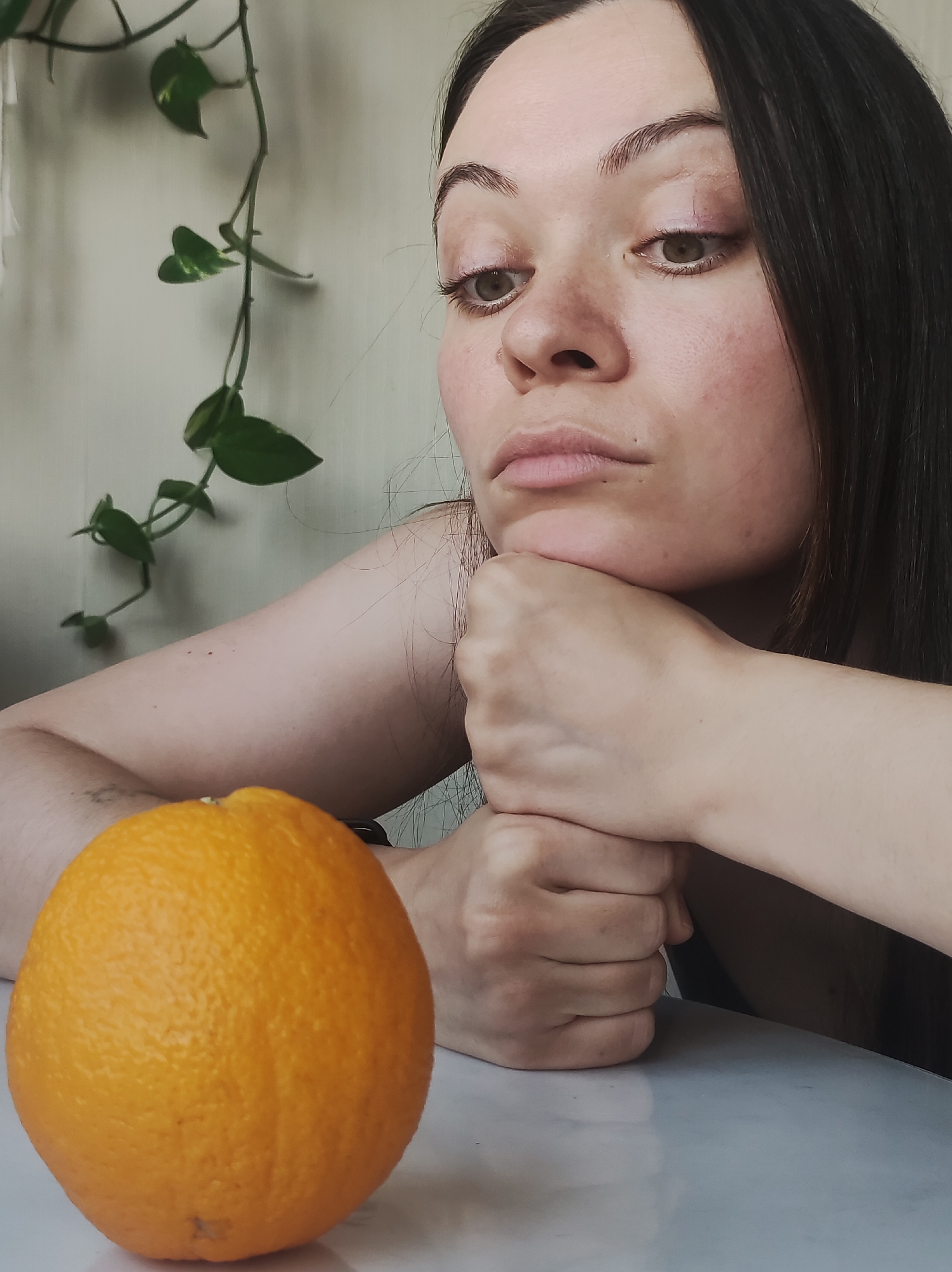 A medium close-up shot captures a young woman in a contemplative pose, looking downwards and to her right with a pensive or slightly melancholic expression. She has dark brown hair, fair skin with visible freckles, and prominent dark eyebrows. Her eyes are a light brown or hazel, and she wears faint pink eyeshadow. Her chin is resting on her clasped hands, which are positioned on a light, reflective surface.
In the lower-left foreground, a single, large, unpeeled orange with a textured rind sits prominently on the same surface. The background features a light-colored wall, against which a vining plant with green, heart-shaped leaves trails downwards. The lighting is soft and natural, indicating daytime. The scene evokes a quiet, reflective atmosphere in an indoor setting in Chaikovsky, Russia. There is no visible text.