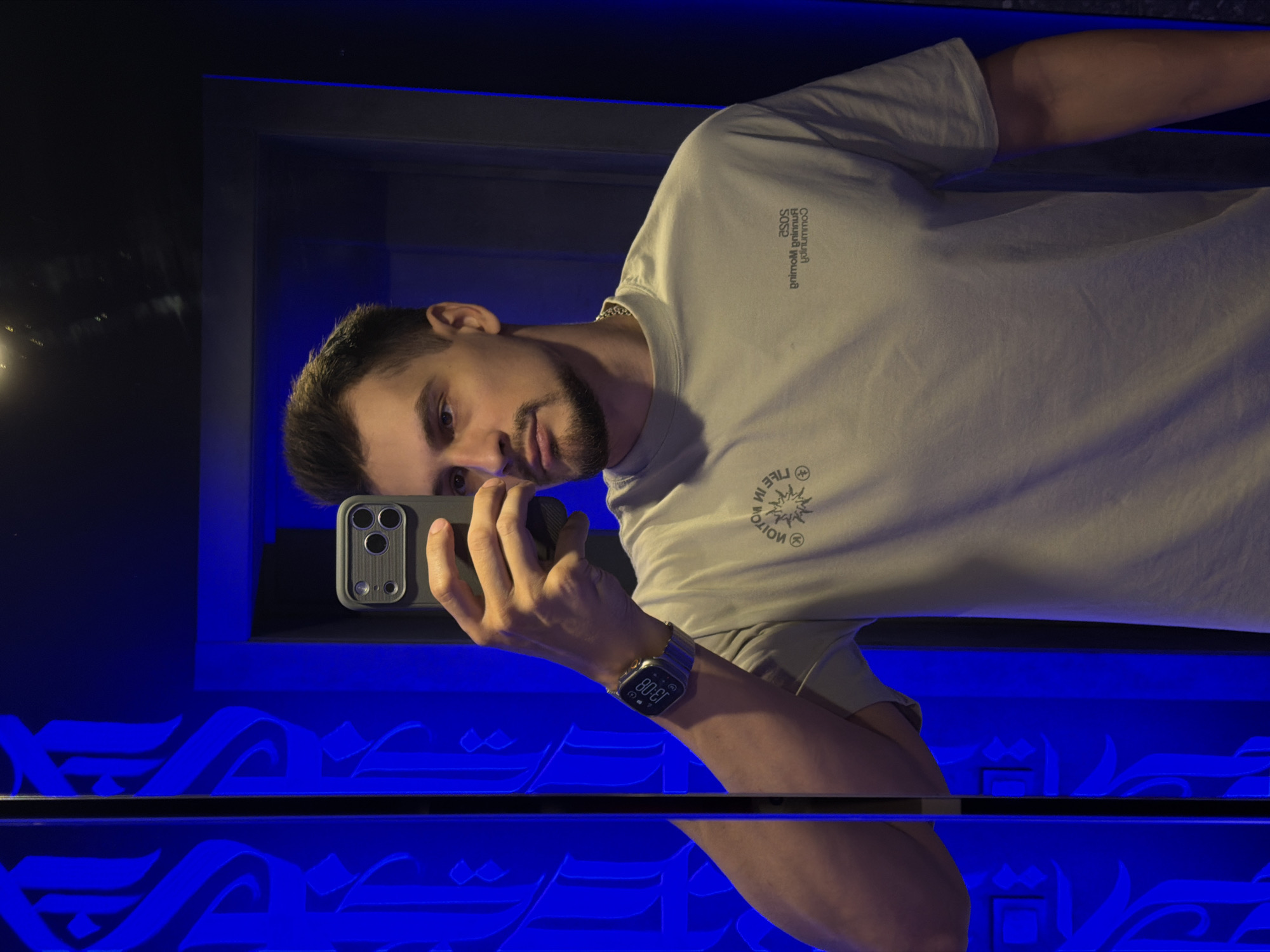The image displays a mirror selfie of a young man, presented in a landscape orientation but originally taken vertically. The man, with short dark hair, a neatly trimmed beard, and mustache, looks directly at the camera with a neutral expression. He is wearing a light-colored (possibly beige or off-white) short-sleeved T-shirt. On his left wrist, he wears a smartwatch that displays the time as "13:08" and various app icons. He holds a smartphone in a grey protective case to take the picture.
The setting appears to be indoors, characterized by dark surroundings and prominent blue illumination. In the background, a horizontal decorative element features intricate, illuminated blue script, resembling Arabic or a similar Middle Eastern calligraphy style, which creates a pattern. This design is also reflected below, suggesting a reflective surface or a repeated motif. The blue lighting creates an atmospheric and modern ambiance.
Visible text on his T-shirt (reversed due to the mirror) includes "SOSE" (likely "ASOS"), "gninurC gninmuC" (likely "Community Running"), and a circular logo with text "3FIL NI WW" (likely "WW IN LIFE") along with a sunburst graphic. The time on his smartwatch is clearly visible as 1:08 PM. The overall scene suggests a contemporary interior, possibly a lounge, restaurant, or modern public space in Krasnoyarsk, Russia.