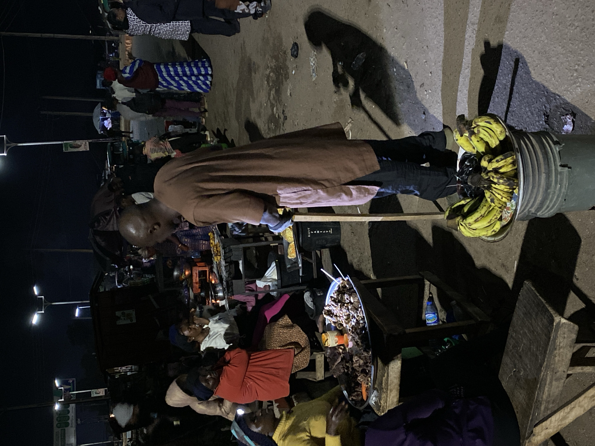 This nighttime image depicts a bustling street market in Bukuru, Nigeria, illuminated primarily by overhead streetlights and supplementary vendor lighting. In the foreground, a dark-skinned man in a brown tunic and dark trousers stands, holding a light wooden stick in his left hand, with his back partially turned to the viewer. To his left, a dark-skinned woman wearing a yellow top and a head covering with a small headlamp is seated behind a large metal tray filled with dark, cooked meat pieces and orange garnishes, accompanied by two white skewers. Adjacent to her, two other dark-skinned women are seated on low wooden stools; one wears a red jacket over a patterned dress, and the other a white long-sleeved top. Below one of these stools, a blue plastic water bottle is visible. A metal basin filled with yellow bananas sits on the unpaved, dusty ground near the standing man's feet. In the midground, additional informal food stalls feature items like grilled food on skewers, and a black "HAPPY FAMILY" branded bin is present. The background shows more individuals walking or congregating, along with various makeshift kiosks and a street banner displaying a portrait. The scene is characterized by deep shadows and bright illumination points, indicative of active commerce in an informal setting after dark.