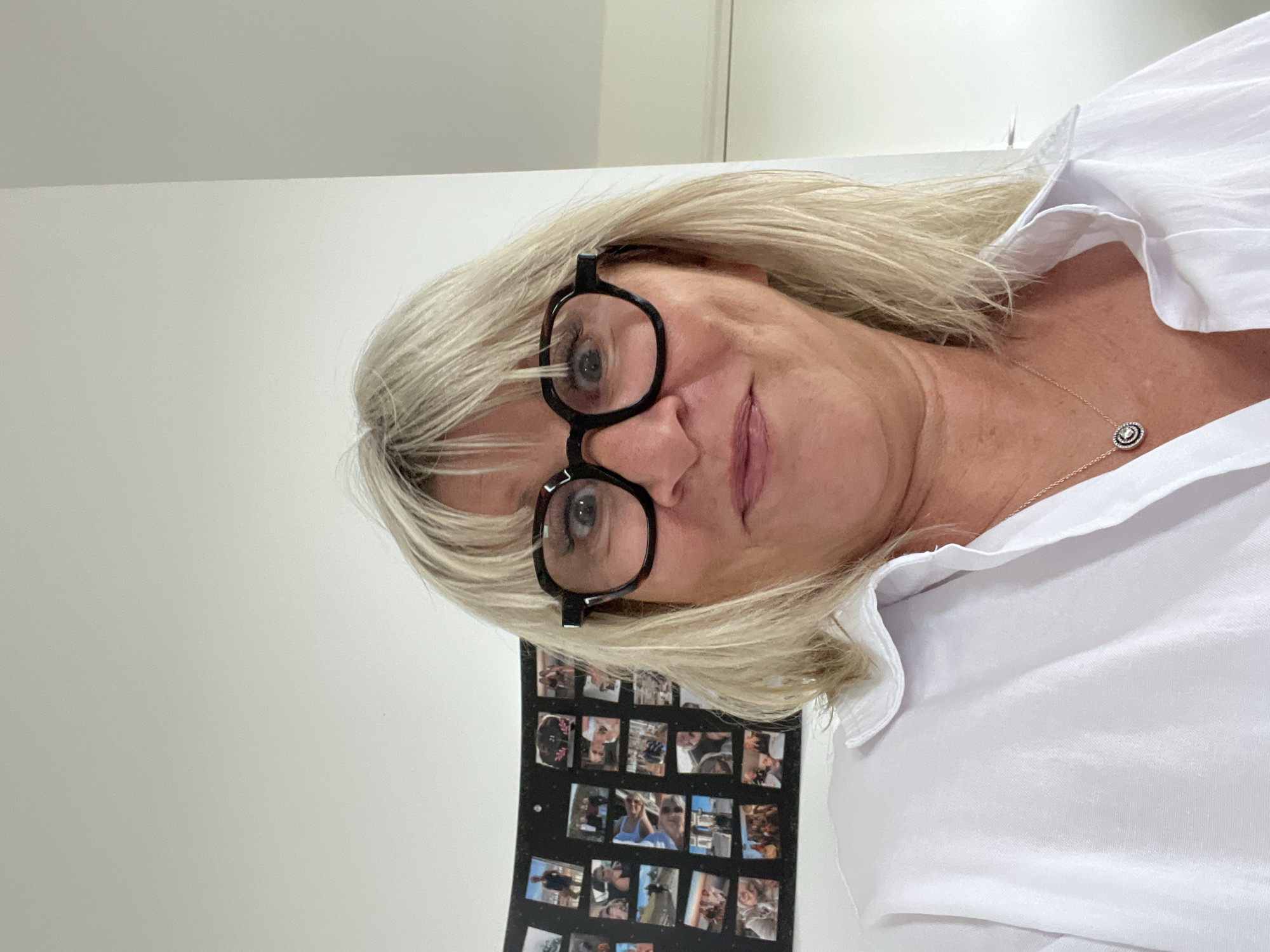 A woman with blonde hair and black-rimmed glasses is the central subject, wearing a white collared shirt and a delicate necklace. She is positioned at an angle to the camera, seemingly indoors with a white wall as the background. A rectangular object, possibly a photo collage, is mounted on the wall behind her. There is no indication that the photo was taken in Chamboeuf, France.