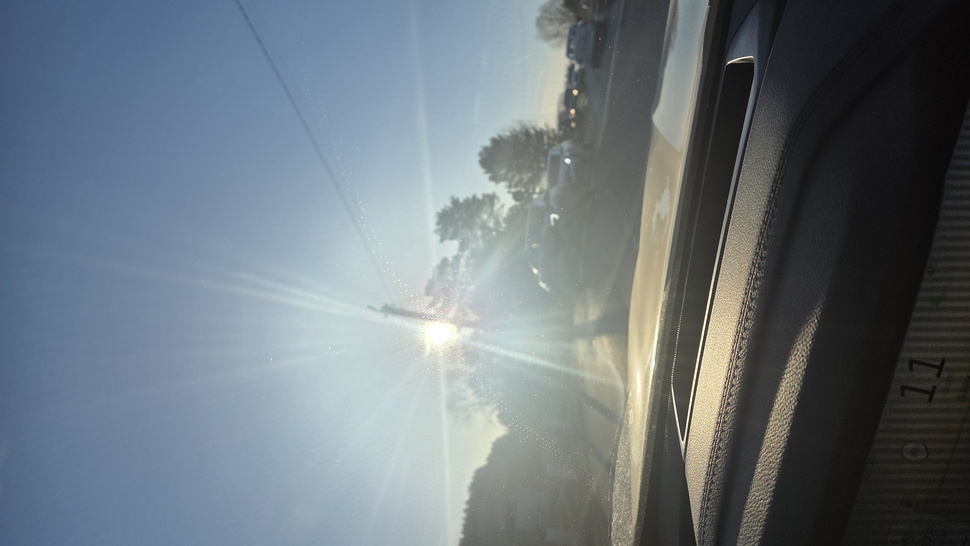 The media file displays a sun-drenched outdoor scene viewed from within a vehicle, likely in Chaikovsky, Russia. The image is currently rotated 90 degrees counter-clockwise.
If viewed in its intended landscape orientation, the scene depicts a brilliant, low-angle sun dominating the lower-middle section, casting intense starburst flares and glare across a moderately dusty or smudged windshield. Beyond the windshield, a clear, light blue sky stretches across the upper left portion of the frame. Below the horizon, a row of bare-branched trees suggests a colder season such as late autumn or winter. A thin, dark line, possibly a utility wire or pole, cuts across the sky. In the mid-distance, several vehicles can be seen, appearing to be either in a line of traffic or parked alongside a road, with sunlight glinting off their surfaces.
The right side of the image reveals the dark, textured interior of the car, likely a door panel or dashboard, characterized by visible stitching and a handle-like recess. This interior section is largely in shadow, providing a strong contrast to the bright exterior. Faint white numbering, specifically "011" or "O11", is discernible on the lower right corner of this dark panel. The intense glare from the low sun indicates either early morning or late afternoon. The overall impression is one of driving or being stationary in a car during a very bright, clear day, facing directly into the sun.