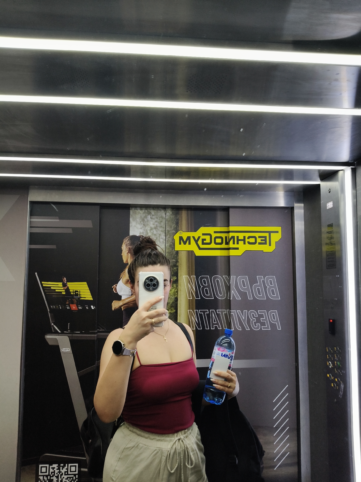 A young woman is taking a mirror selfie in what appears to be an elevator or a gym area with reflective surfaces. She is wearing a burgundy crop top and light-colored pants. She holds a smartphone in her right hand, obscuring part of her face, and a blue plastic water bottle in her left hand. A smartwatch is visible on her left wrist. Behind her, a large poster displays fitness-related imagery and text, including the "TECHNOGYM" logo. The environment is modern and well-lit, with metallic finishes and bright strip lights.