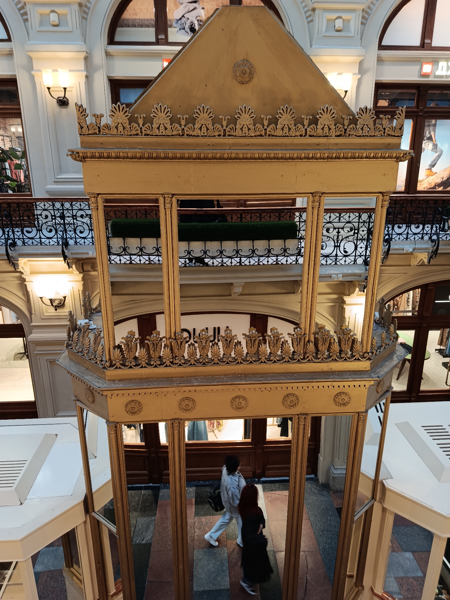 This image depicts the interior of a grand, multi-story building, likely a department store or shopping gallery in Moscow, Russia. The central feature is a prominent, ornate golden structure, possibly an elevator or a decorative enclosure, richly adorned with classical motifs including rosettes, acanthus leaves, and palmettes. This structure has multiple tiers, with the upper section featuring open frameworks and intricate decorative balustrades.
Two people are visible at the base of this golden structure, walking on a patterned tiled floor. One person wears light-colored clothing, and the other is dressed in dark attire, suggesting they are shoppers or visitors. The building's interior showcases a sophisticated architectural style with arched walkways, high ceilings, and multiple levels. Each level is bordered by elaborate black wrought-iron railings, adding to the opulent aesthetic.
Wall sconces emit warm light, illuminating the cream-colored walls and the detailed architectural elements. Shop fronts with display windows are visible on the upper levels, one featuring a large poster. Artificial greenery is discernible behind some of the railings on the upper floors. Partially visible Cyrillic text in a shop window on an upper level reads "ДУХИ" (DUHI), meaning "perfumes" or "spirits." The scene conveys a sense of historical grandeur and elegance within a commercial setting.
