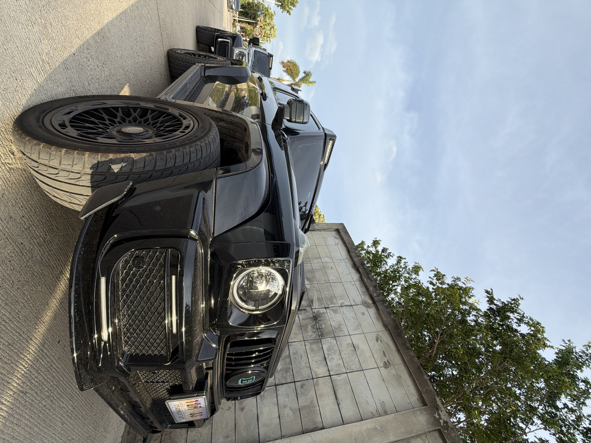 Two black Mercedes-Benz G-Wagons are parked on a concrete street next to a textured concrete wall with trees and sky visible in the background. The foremost G-Wagon is black with a distinctive grill and black aftermarket rims. A license plate is visible on the front of the car. The second G-Wagon, parked behind the first, is silver. The image appears to have been taken in Ko Samui District, Thailand, given the presence of palm trees and the style of the license plate.