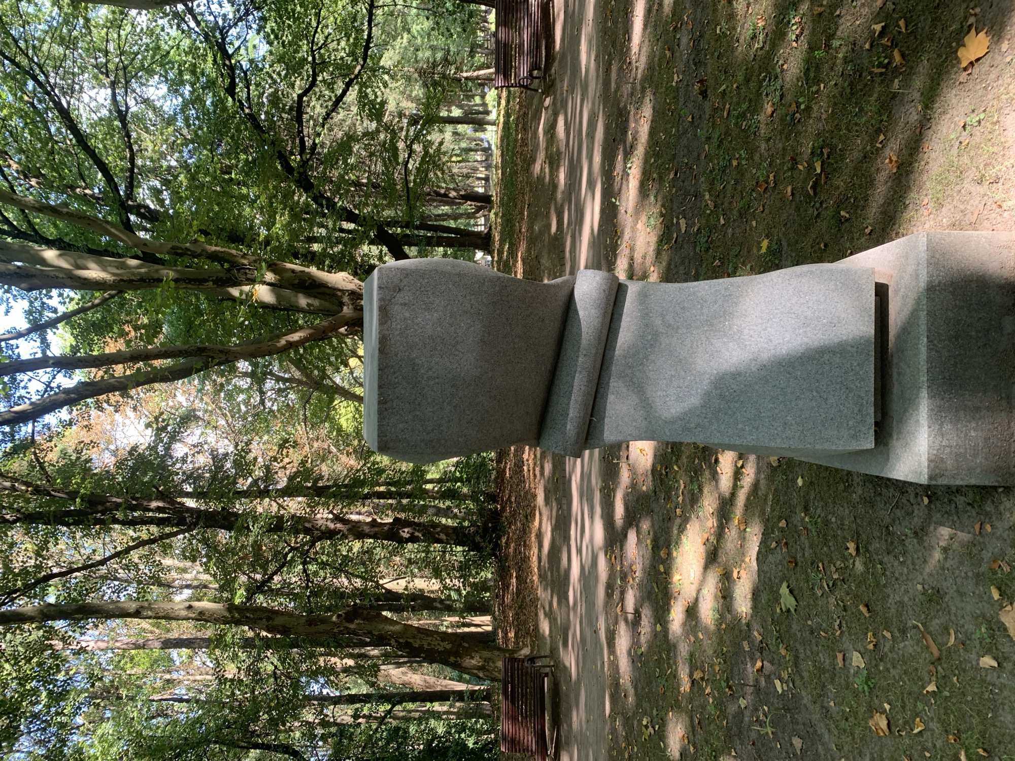 The image depicts a park scene in Lviv, Ukraine. In the foreground, a tall, grey stone sculpture with rounded, abstract forms is positioned on a tiered base. The sculpture has a rough, textured surface. Behind the sculpture, a pathway curves through a park with numerous tall trees casting dappled sunlight and shadows on the ground, which is covered with fallen leaves. Two empty, dark red wooden benches are visible along the pathway in the middle ground, one to the left and one to the right of the frame. The overall environment suggests an autumn day in a tranquil urban park.