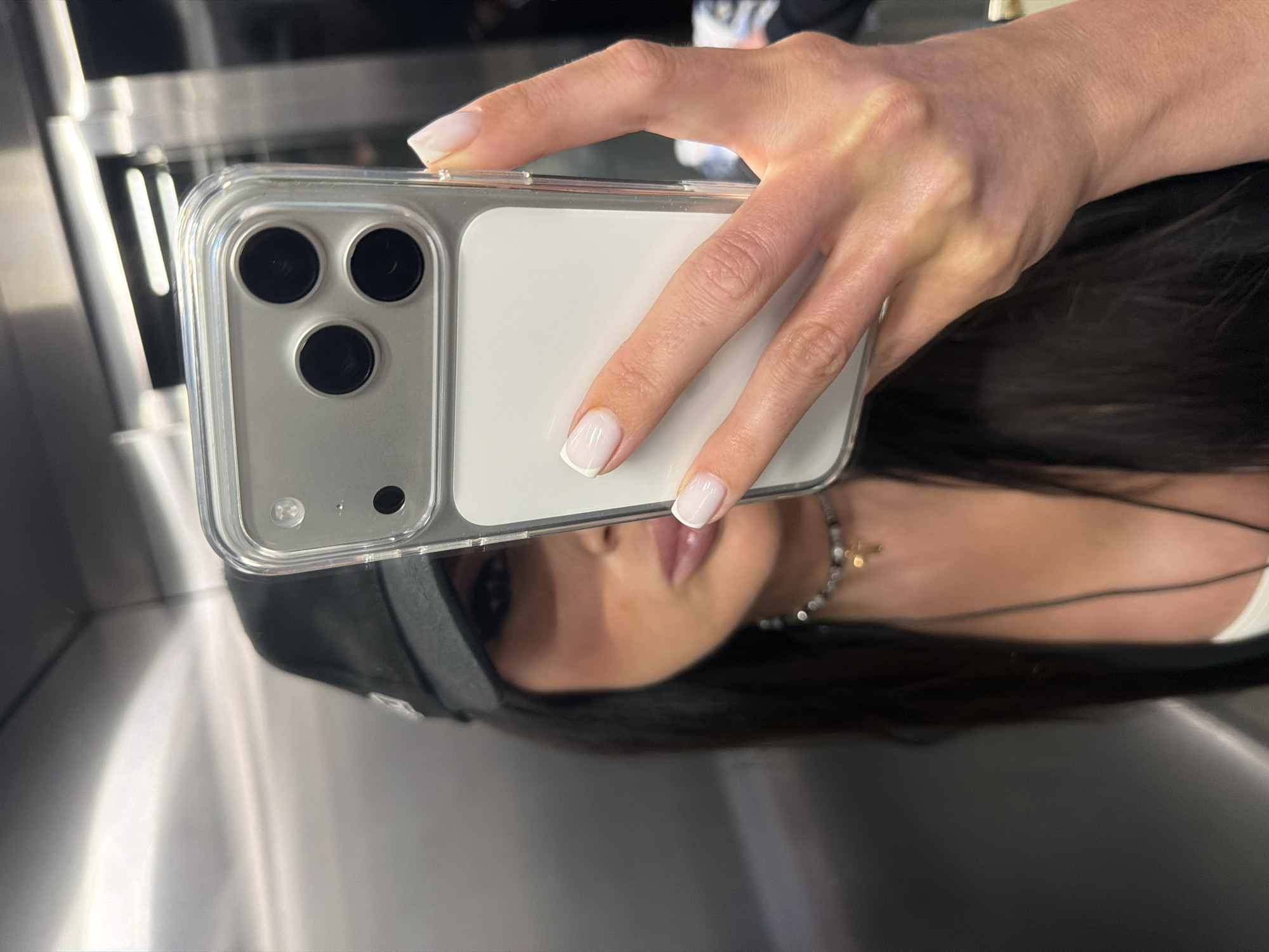A close-up, horizontally oriented shot depicts a person holding a light-colored smartphone with a clear protective case, capturing their partial reflection. The hand, with neatly manicured nails featuring white French tips, grips the phone, which displays a triple camera array and a flash unit on its upper left side. The phone's back is white or silver.
The reflective surface on which the image is captured reveals an inverted and slightly distorted reflection of the person's face, neck, and dark hair. Visible facial features include glossy, plump lips. The person appears to be wearing a dark baseball cap or similar headwear, and a delicate necklace with a small, gold-colored cross pendant is visible around their neck.
The setting, deduced from the blurred reflections in the background, appears to be a modern indoor environment, possibly an elevator or a space with polished metallic walls and bright, artificial lighting. The smooth, shiny surfaces create elongated highlights and soft gradients. The image portrays a casual selfie being taken in such a contemporary setting, located in Boreti, Montenegro. No text is visible.