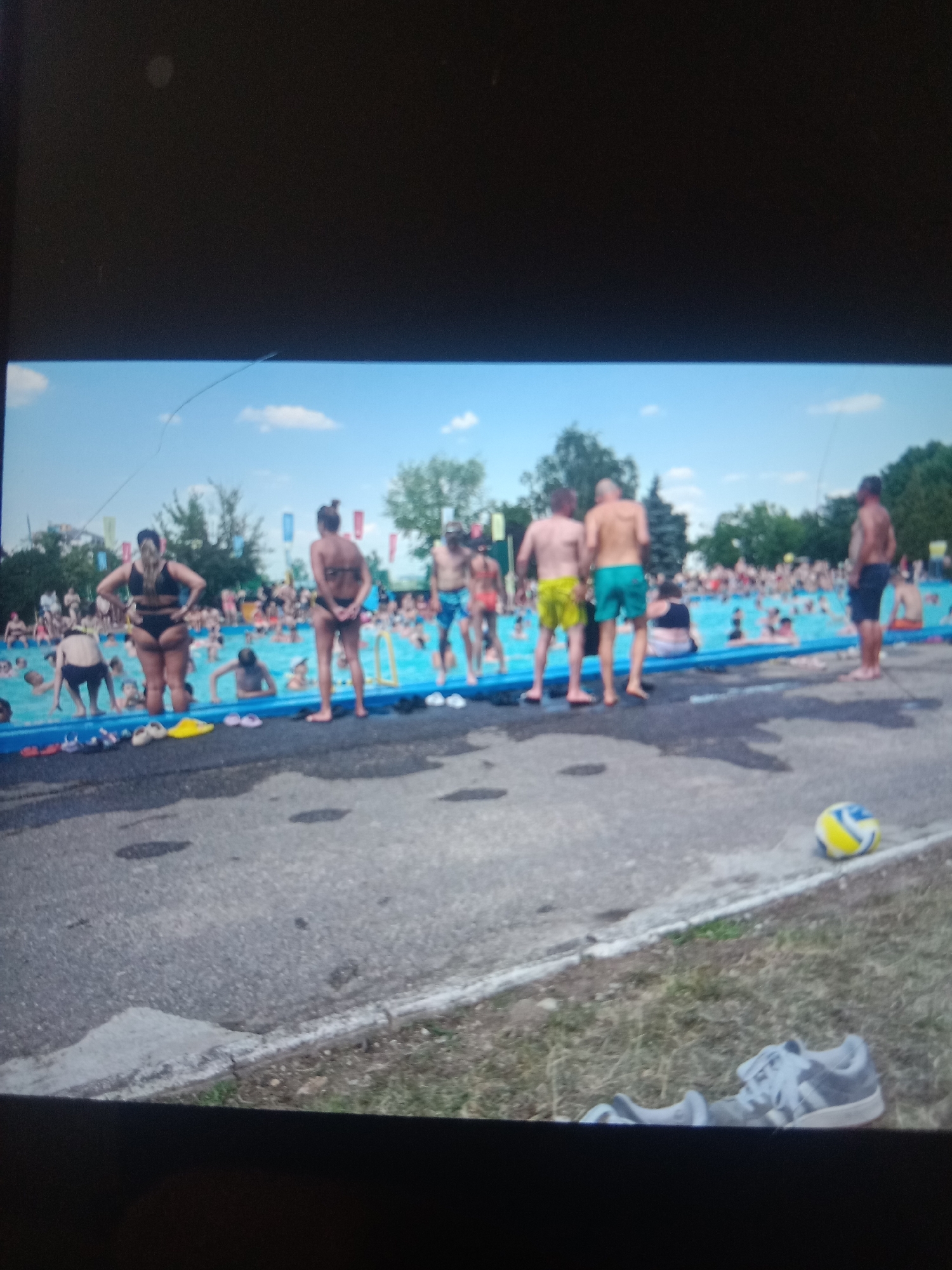 The image depicts a crowded outdoor swimming pool area, likely during warm weather. In the foreground, a paved surface transitions to a grassy area where a single white athletic shoe with grey laces and socks are placed. Further into the scene, a yellow, blue, and white volleyball rests on the grass. The main focus is a large, bright blue swimming pool filled with numerous people, many of whom are partially submerged in the water. Several individuals are visible standing at the edge of the pool, wearing swimwear. The background features trees and flags, suggesting a recreational park or water park setting. The lighting indicates daytime. The image does not contain any specific identifiable landmarks or features that would definitively place the location in the city of Kielce, Poland.