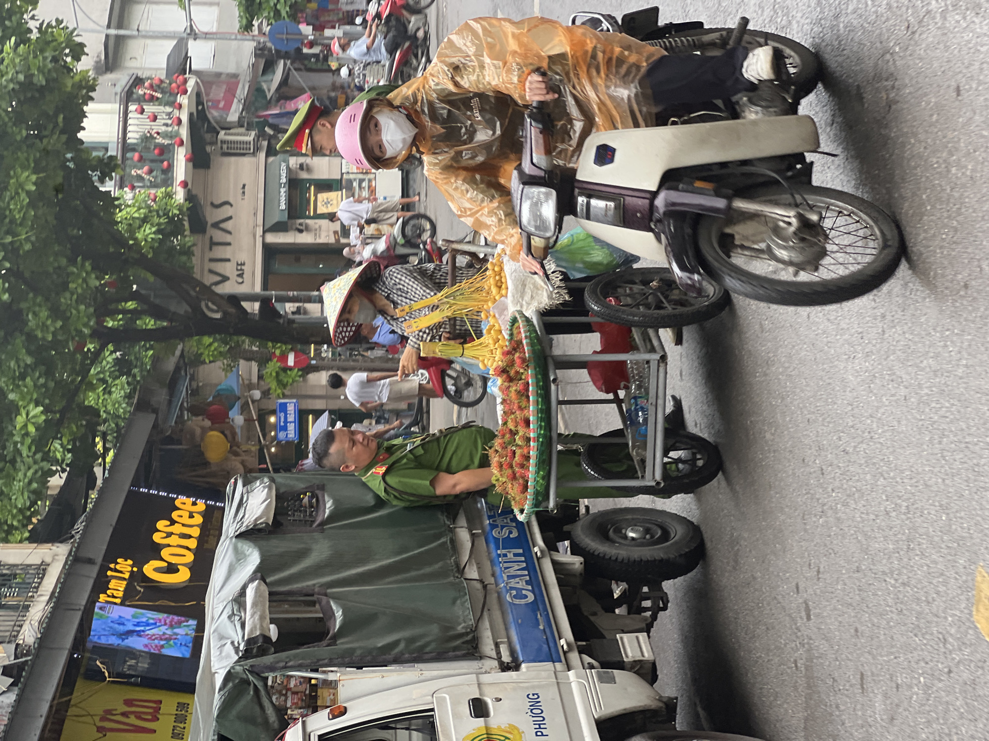 In Hanoi, Vietnam, on a wet asphalt street, a scene of daily life unfolds. A white truck with a green tarp is parked on the left, bearing the blue inscription "CẢNH SÁT" (Police) and "PHƯƠNG" (Ward). Next to it, a man in a green police uniform stands, overseeing a cart filled with rambutan fruit. To the right, a woman on a scooter wears an orange rain poncho and a pink helmet, her face partially obscured by a white mask. She is looking forward, her hands on the handlebars, suggesting she is either stopped or in motion. In the background, a building with a sign reading "VITAS CAFE" is visible, along with other shops and people on foot and on motorcycles. The atmosphere is that of a busy street scene in a Vietnamese city, with vendors, law enforcement, and pedestrians navigating the urban environment.
